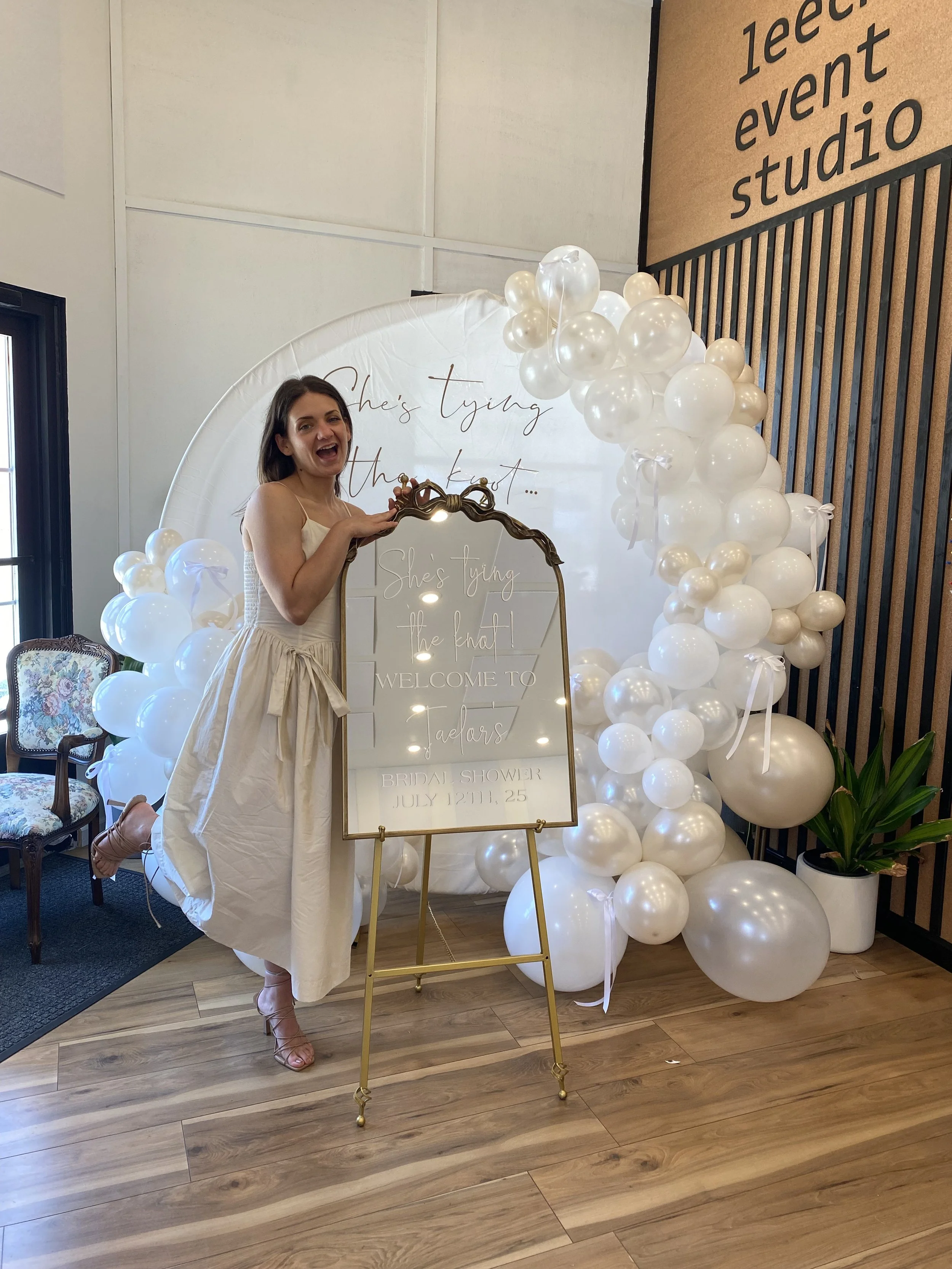 A woman in a white dress standing on one leg, smiling, in front of a decorative backdrop with white balloons, a mirror, and a welcome sign at a bridal shower event.