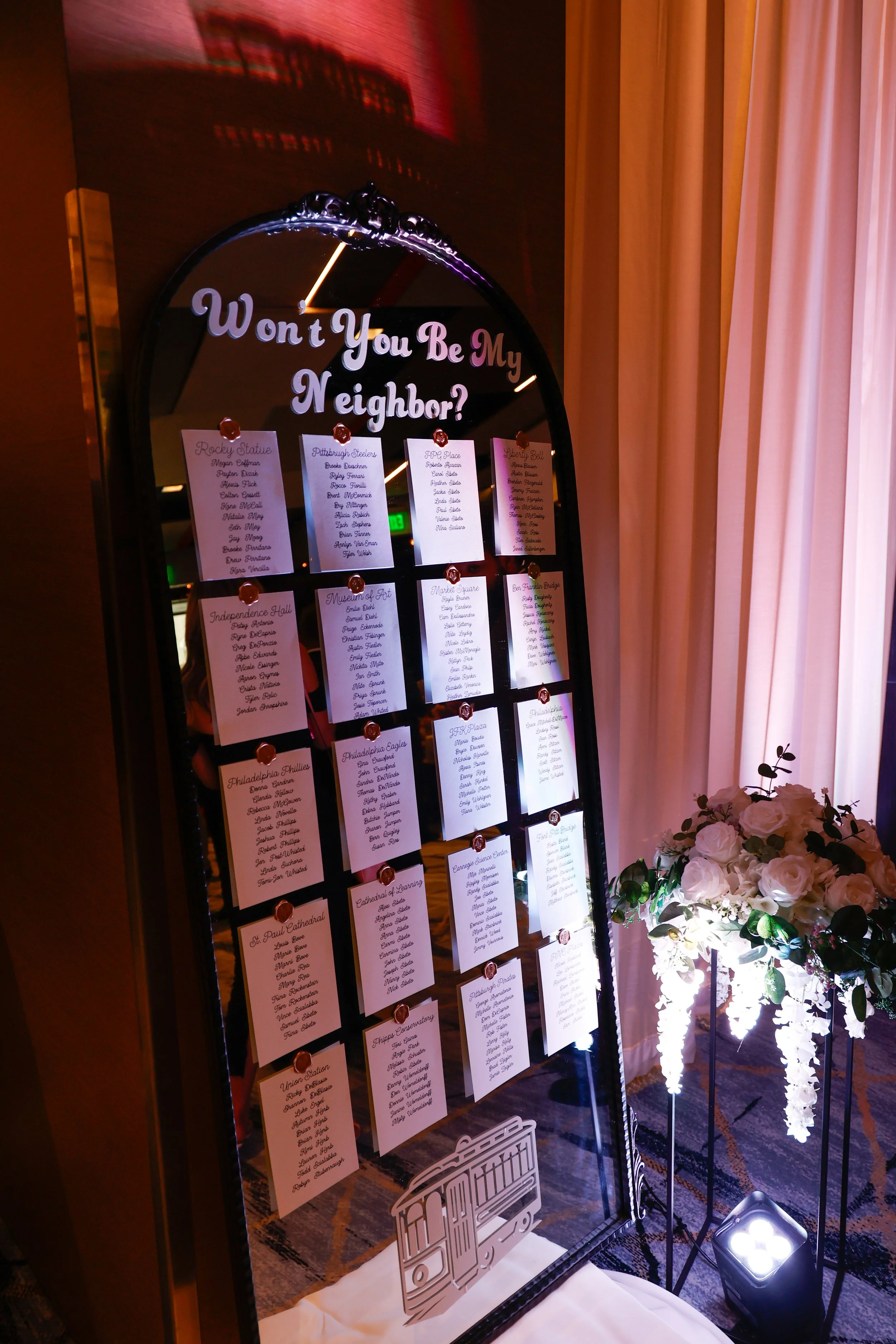 A mirror standing on the floor with a seating chart for an event, titled "Won't You Be My Neighbor?" in pink and purple lettering at the top. The chart lists various locations, each with several names underneath. To the right of the mirror, there is 