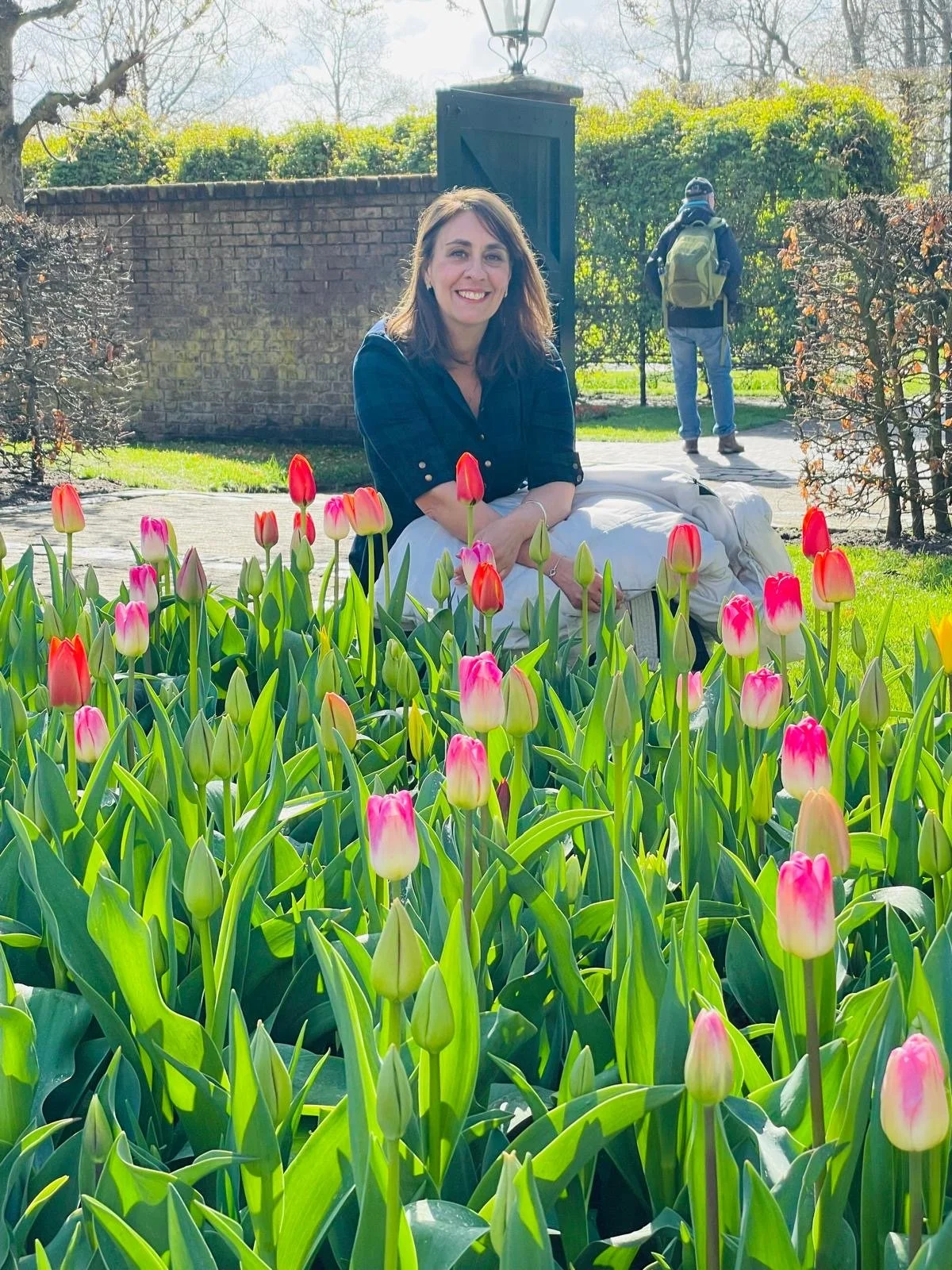 Woman smiling while sitting among pink and white tulips in a garden with bright sunlight and greenery, with a person walking in the background.