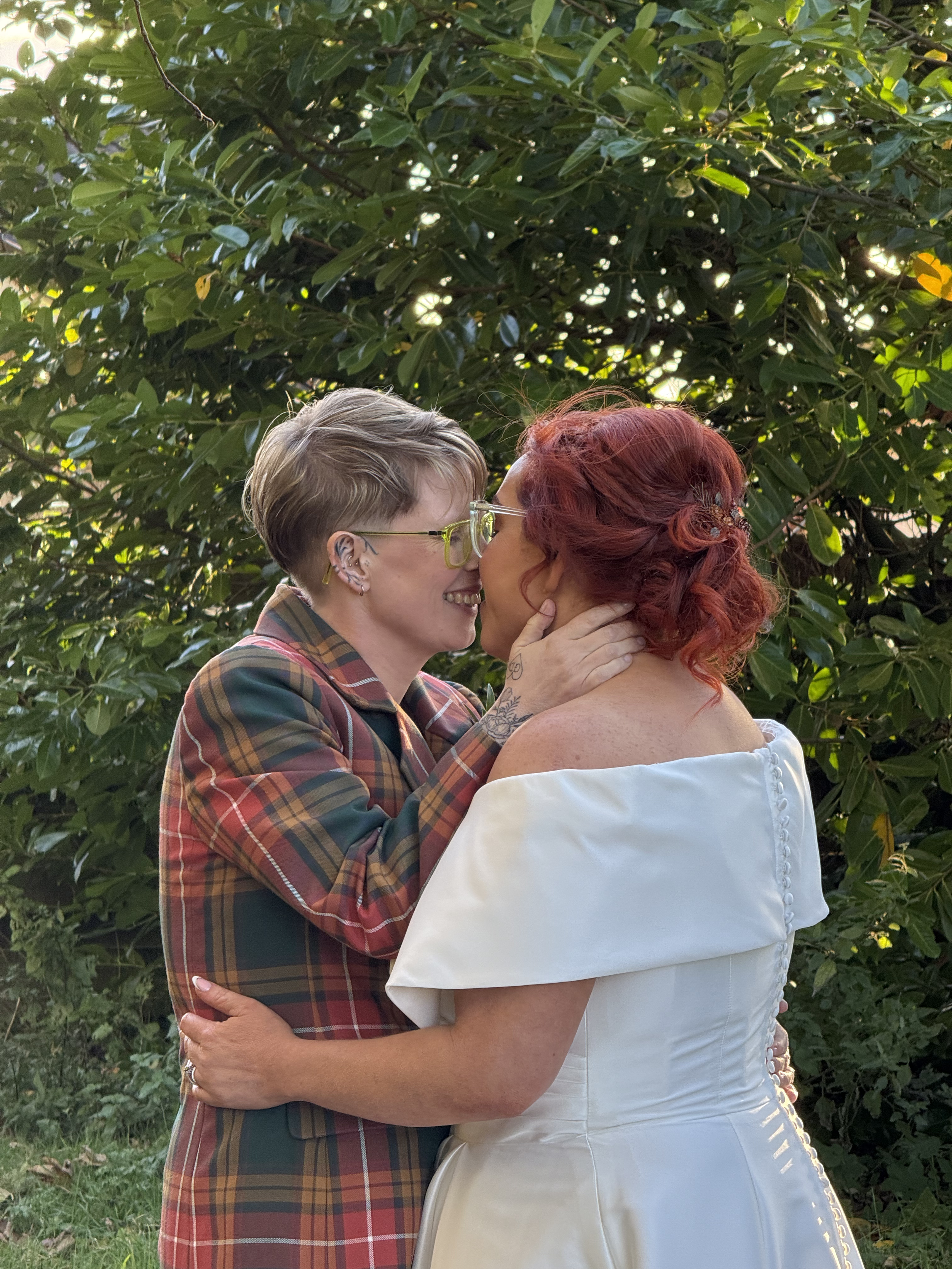 Two women sharing a kiss outdoors, one wearing a plaid blazer and glasses, and the other in a white off-shoulder dress with red hair.