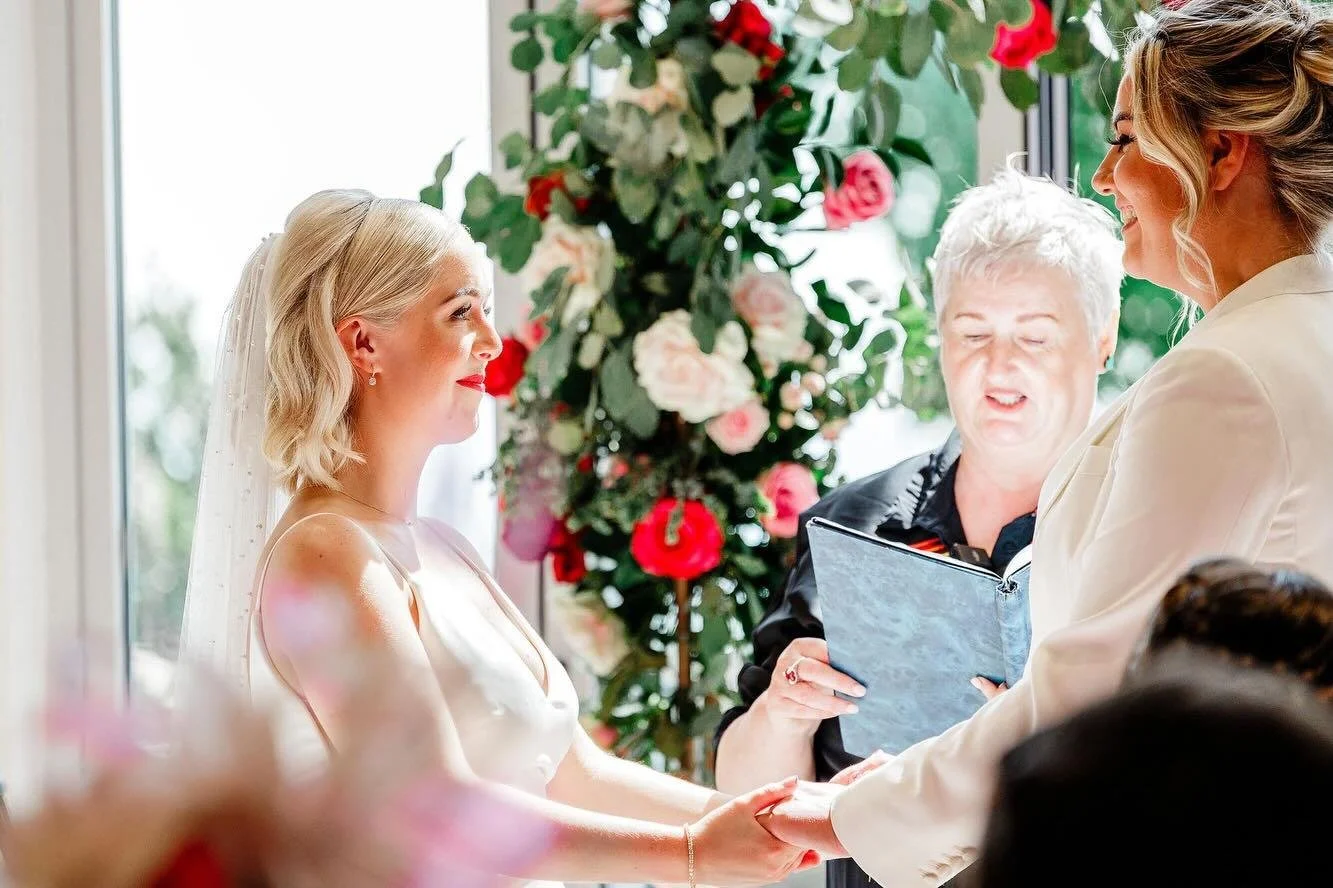 Bride and groom holding hands during wedding ceremony with officiant reading vows; floral arrangements in the background.