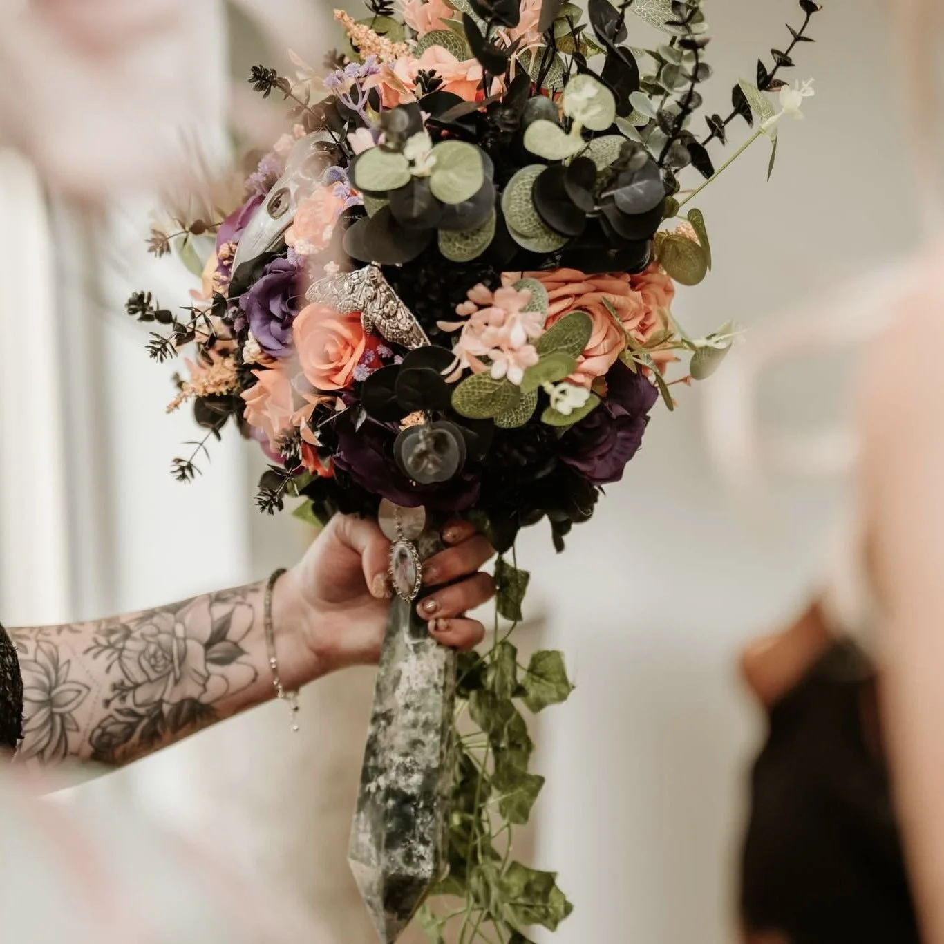 A hand holding a bouquet of pink, purple, and black flowers with green foliage, featuring a butterfly and decorative ribbons.