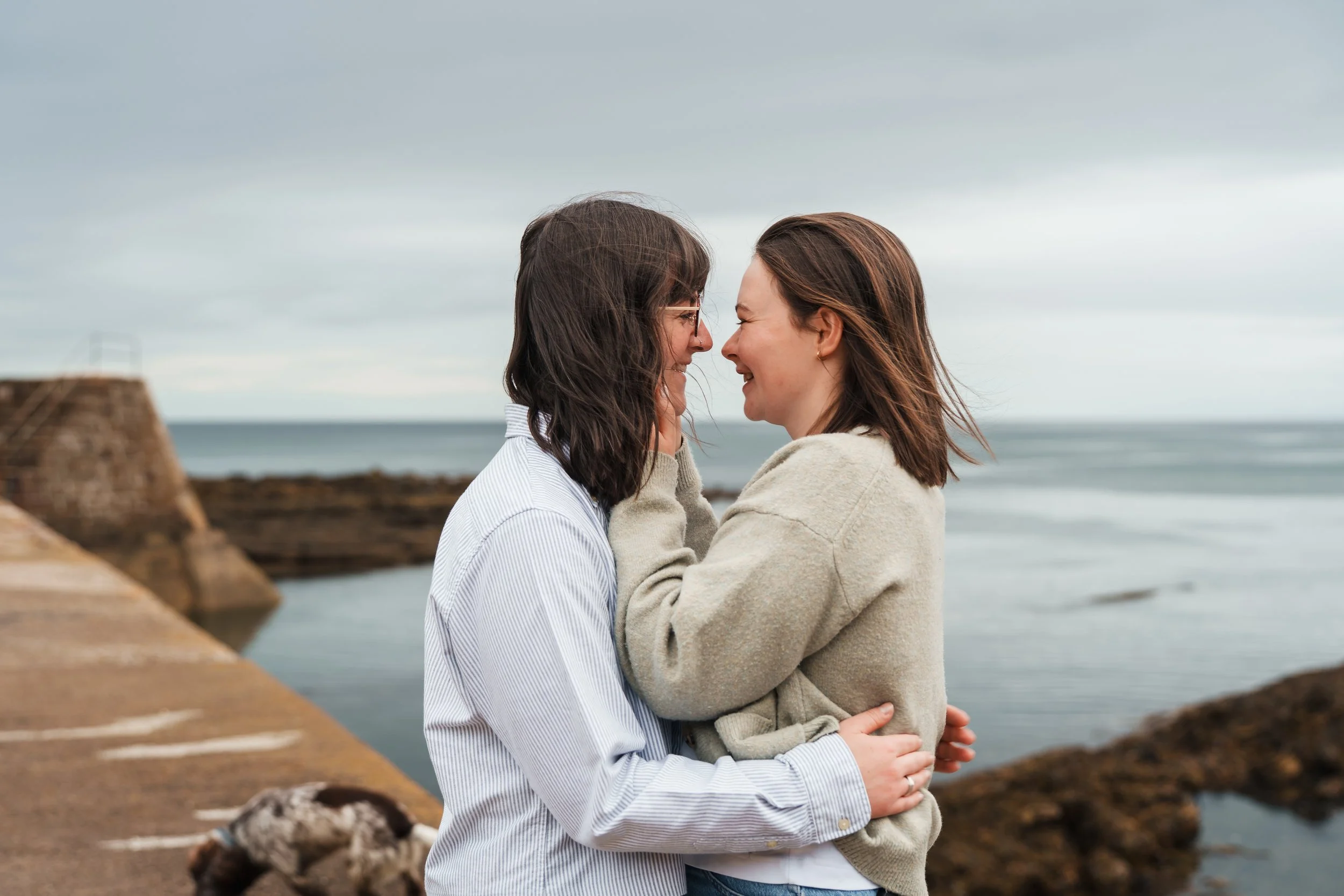 Two women face each other, touching noses and smiling, at the beach with water and rocks in the background.