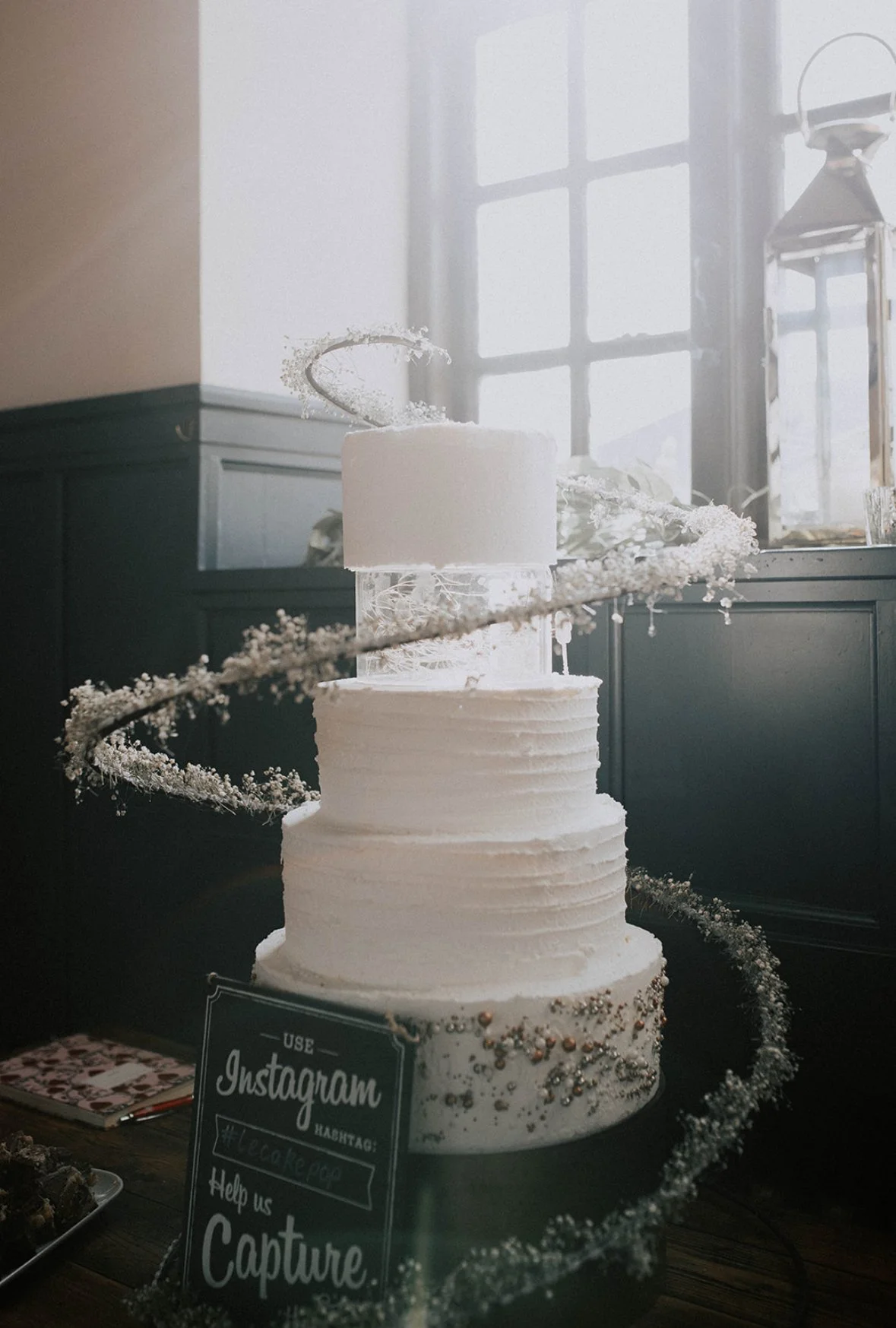 A three-tiered white wedding cake with textured icing and decorative beads at the base, topped with a small modern cake topper, set on a table near a window with natural light.