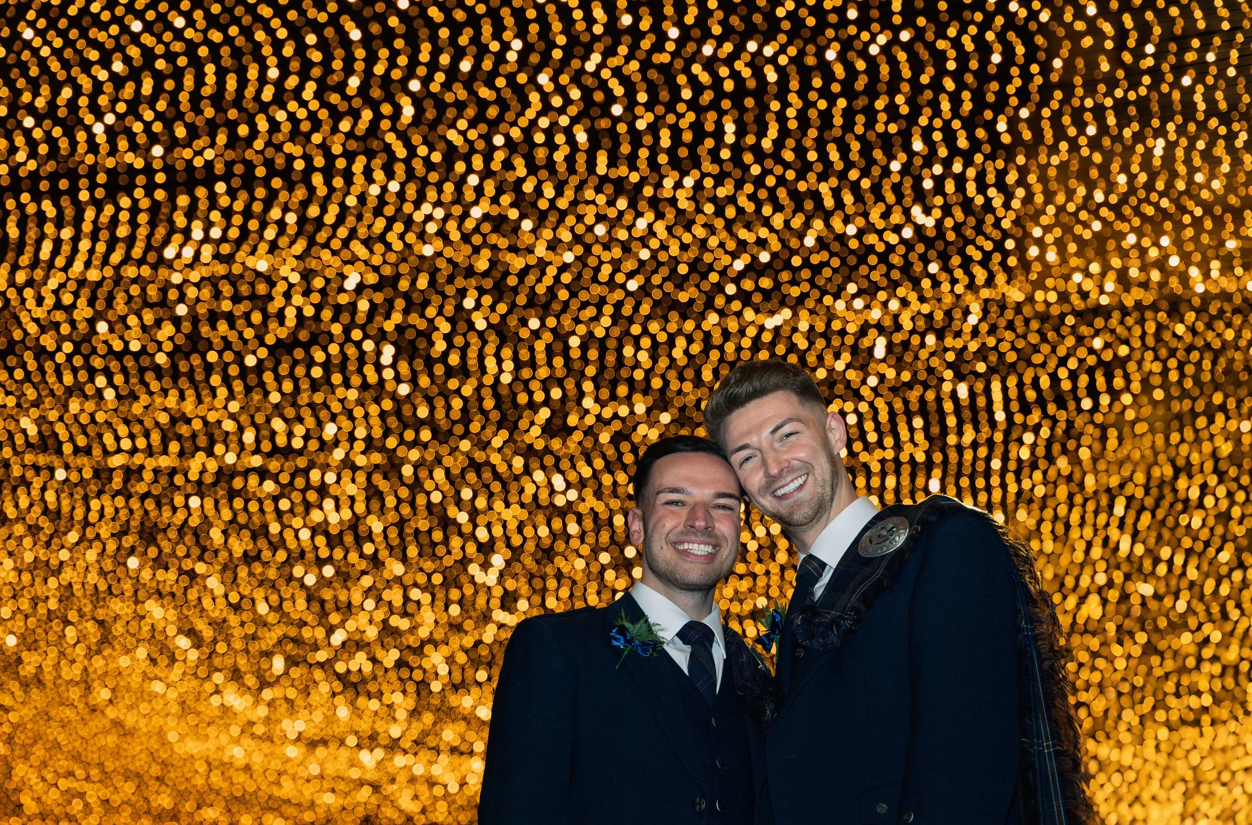 Two men in suits smiling, standing close together with arms around each other's shoulders, under a ceiling decorated with numerous small golden lights.