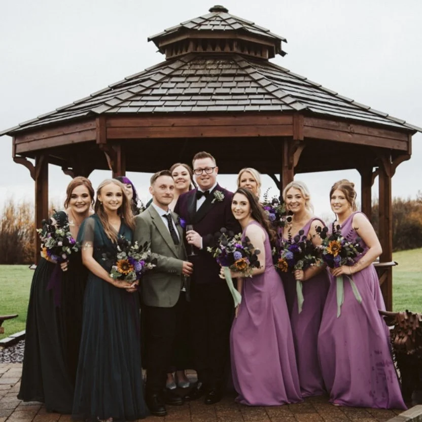 A wedding group photo of nine people, including bride and groom, standing under a wooden gazebo outdoors, with the wedding party holding bouquets of flowers and dressed in formal attire.