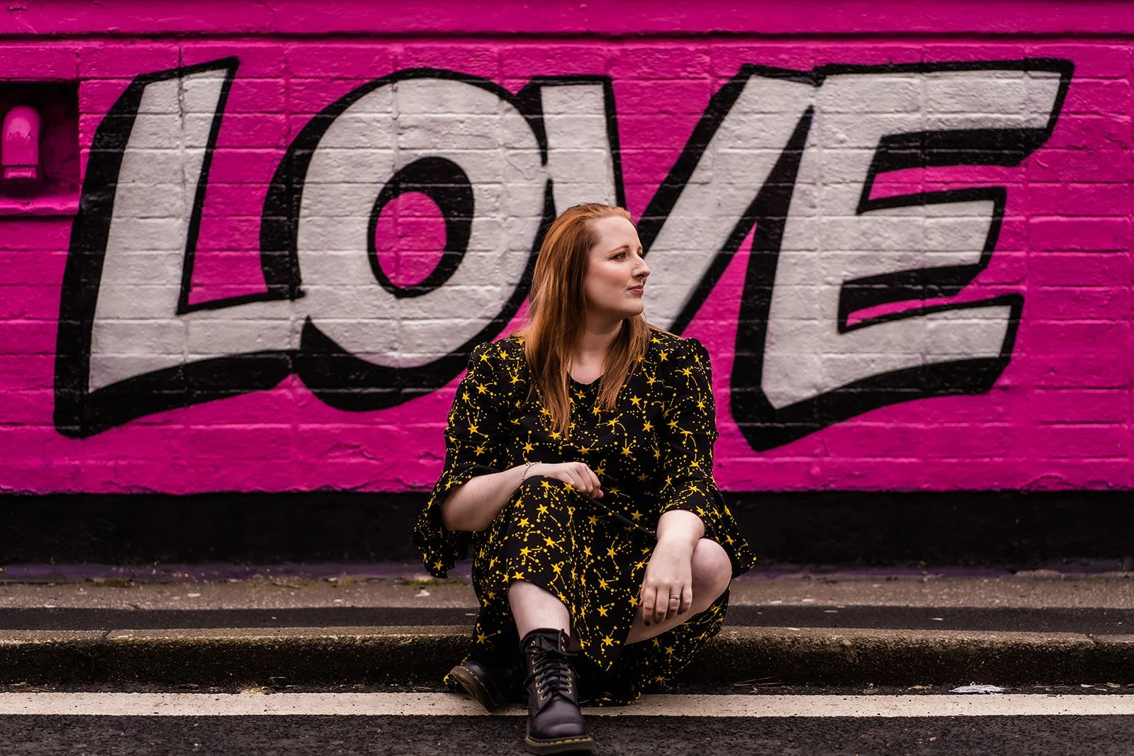 A woman with red hair wearing a black dress with yellow star patterns, sitting on the street curb against a bright pink wall with black and white graffiti spelling 'LOVE.'