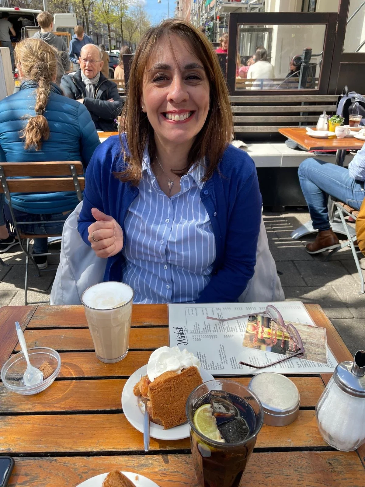 Woman smiling at outdoor cafe table with slice of pumpkin pie, whipped cream, coffee, and soda with lemon, with people sitting and walking in the background.