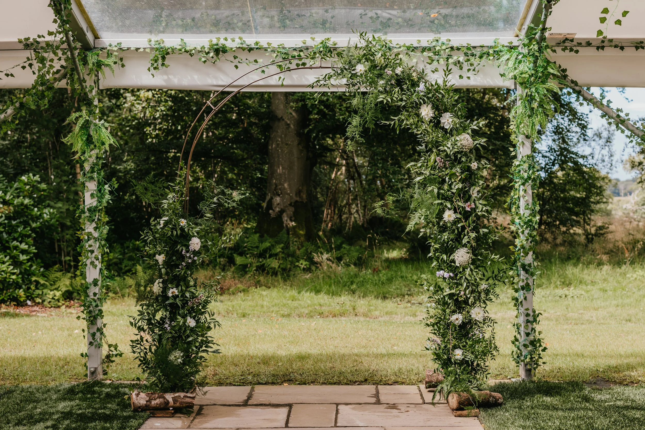 Outdoor wedding arch decorated with green foliage and white flowers on a patio, overlooking a grassy area and trees.