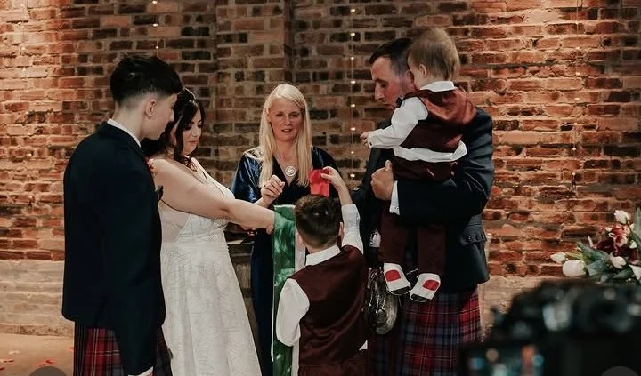 Group of people at a wedding ceremony, with a woman in a white dress and a man in plaid kilt holding a young child, as others participate in a traditional ritual against a brick wall background.