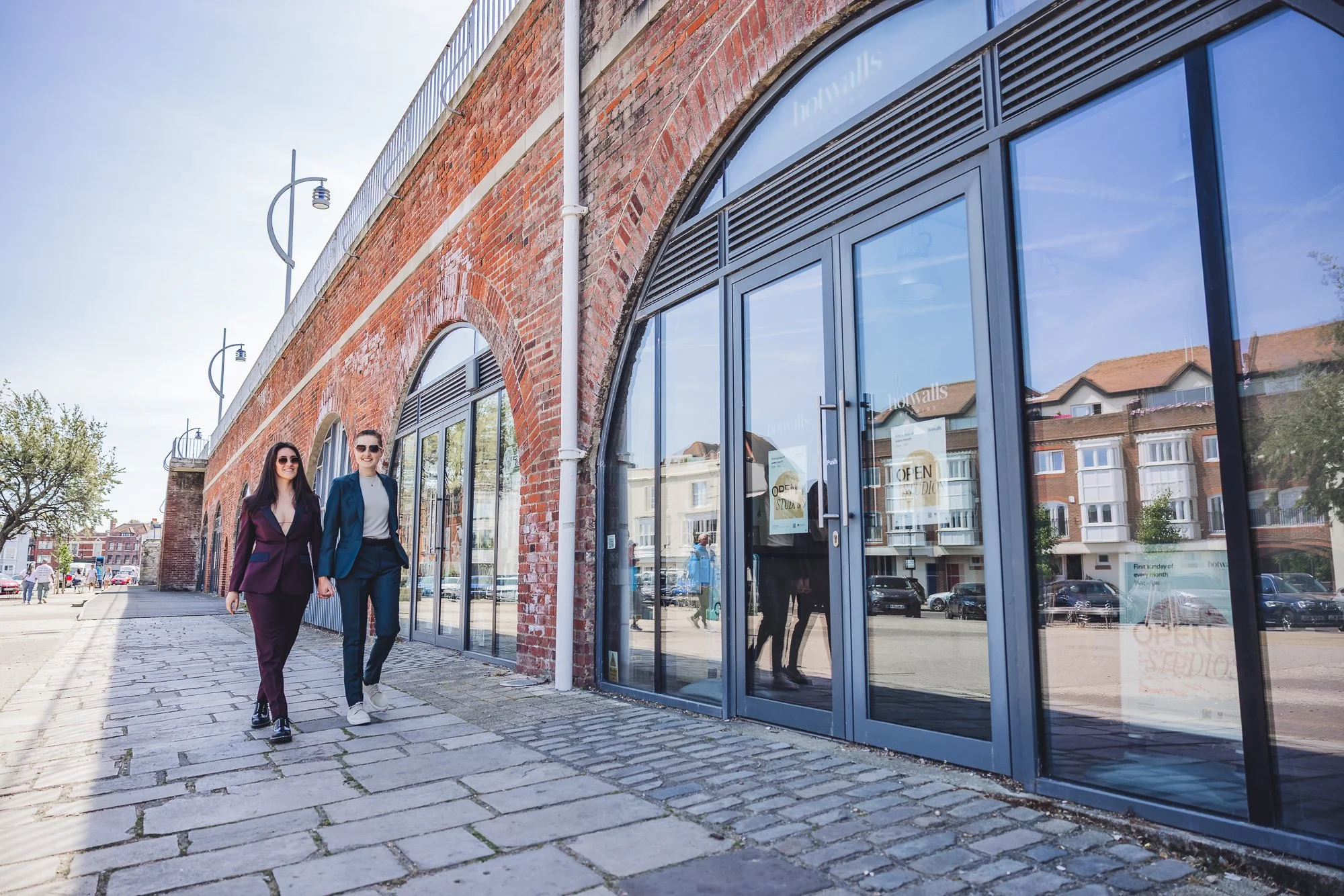 Two women walking on a cobblestone sidewalk next to a modern glass storefront with brick walls in an urban area on a sunny day.