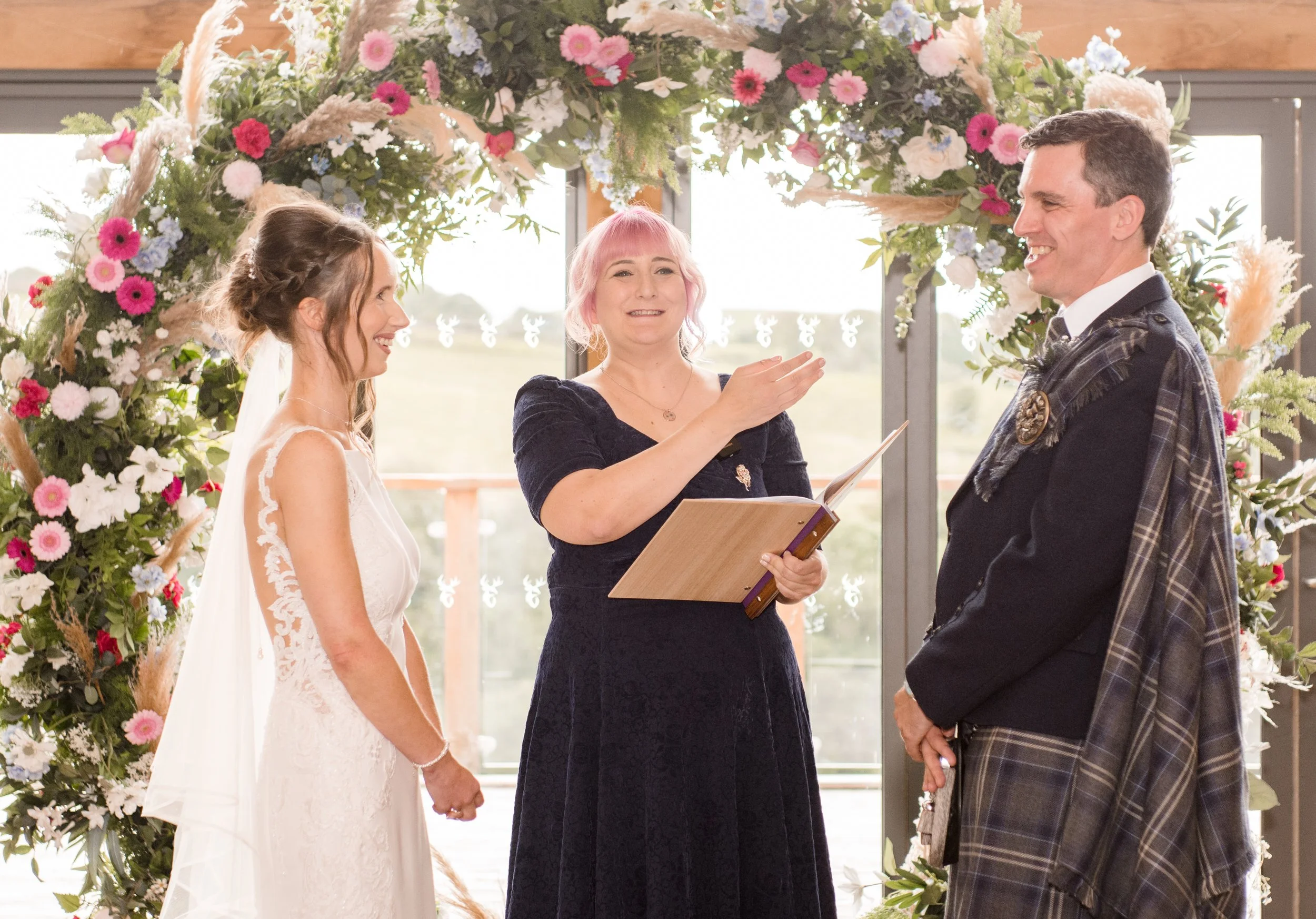 A couple getting married at their wedding ceremony, standing in front of a floral arch with a woman officiating, indoors with large glass windows in the background.