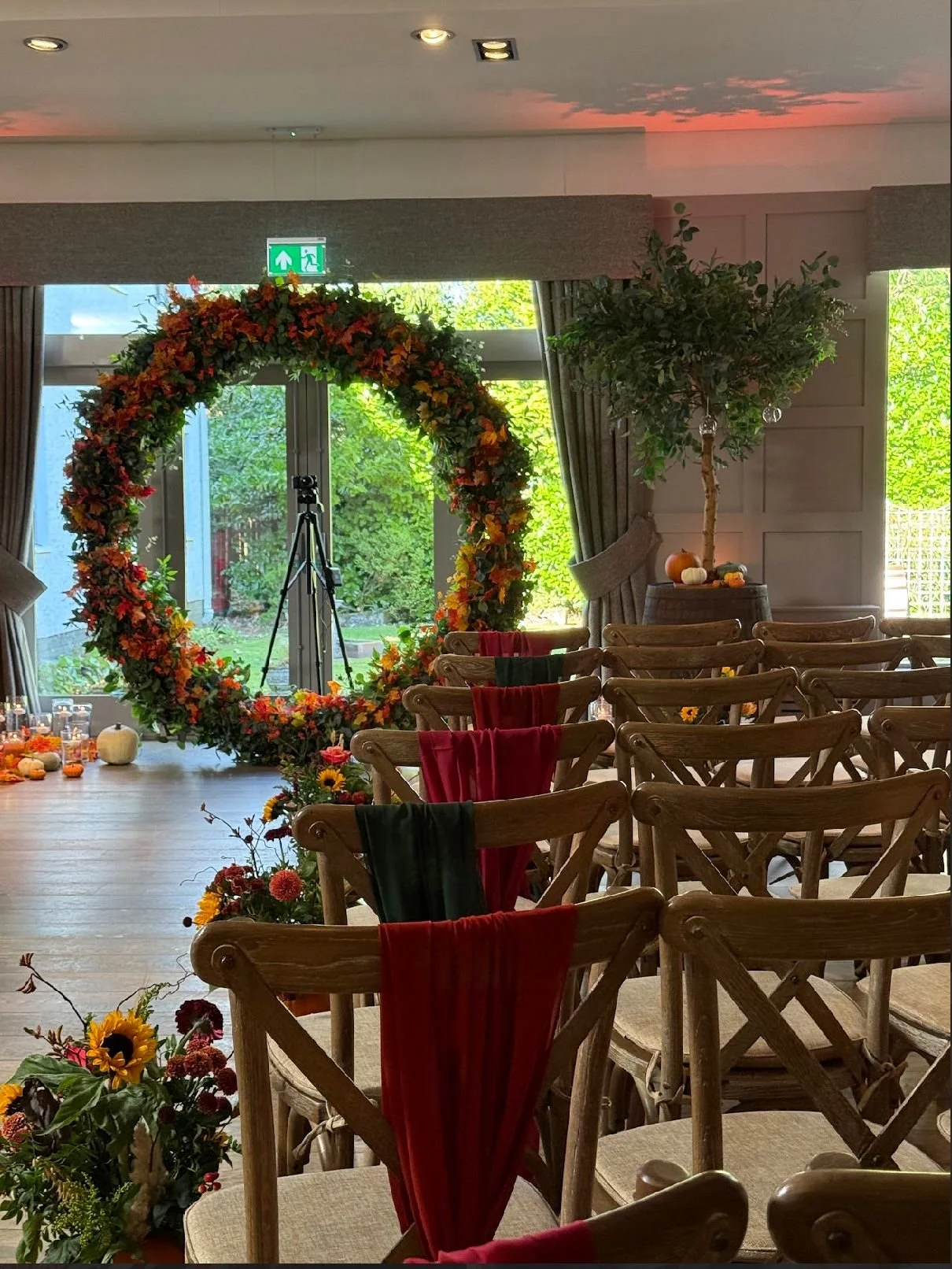 An indoor event space decorated with a large floral wreath, pumpkins, and autumnal decorations. Rows of wooden chairs with red and green cloths attached, facing forward. Large windows with curtains, trees visible outside. A tripod with a camera or device in front of the wreath.