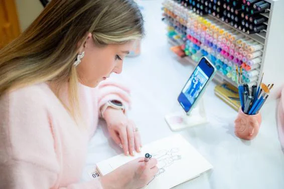 A woman drawing in a sketchbook, with nail polish colors on a rack, a smartphone on a stand, and a cup holding pens and brushes on her desk.