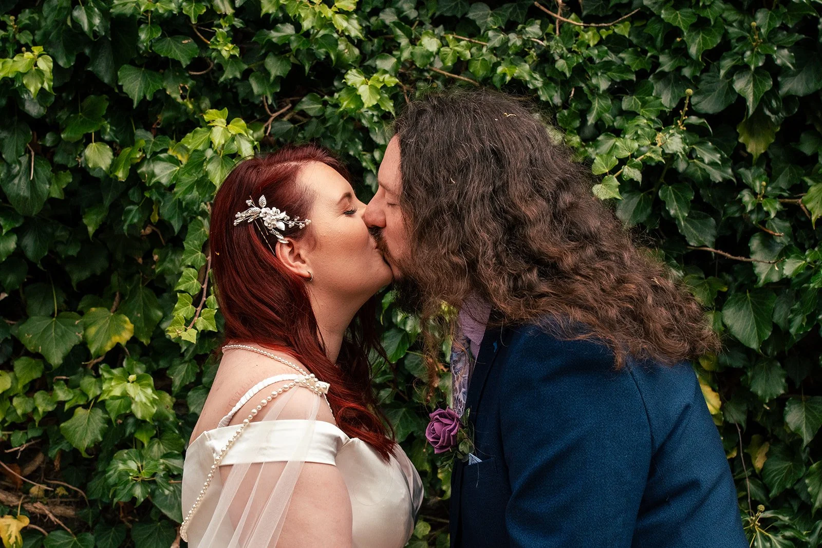 A bride and groom kiss in front of a leafy green background, with the bride wearing a white dress and pearl accessories, and the groom dressed in a dark suit with a purple boutonniere.