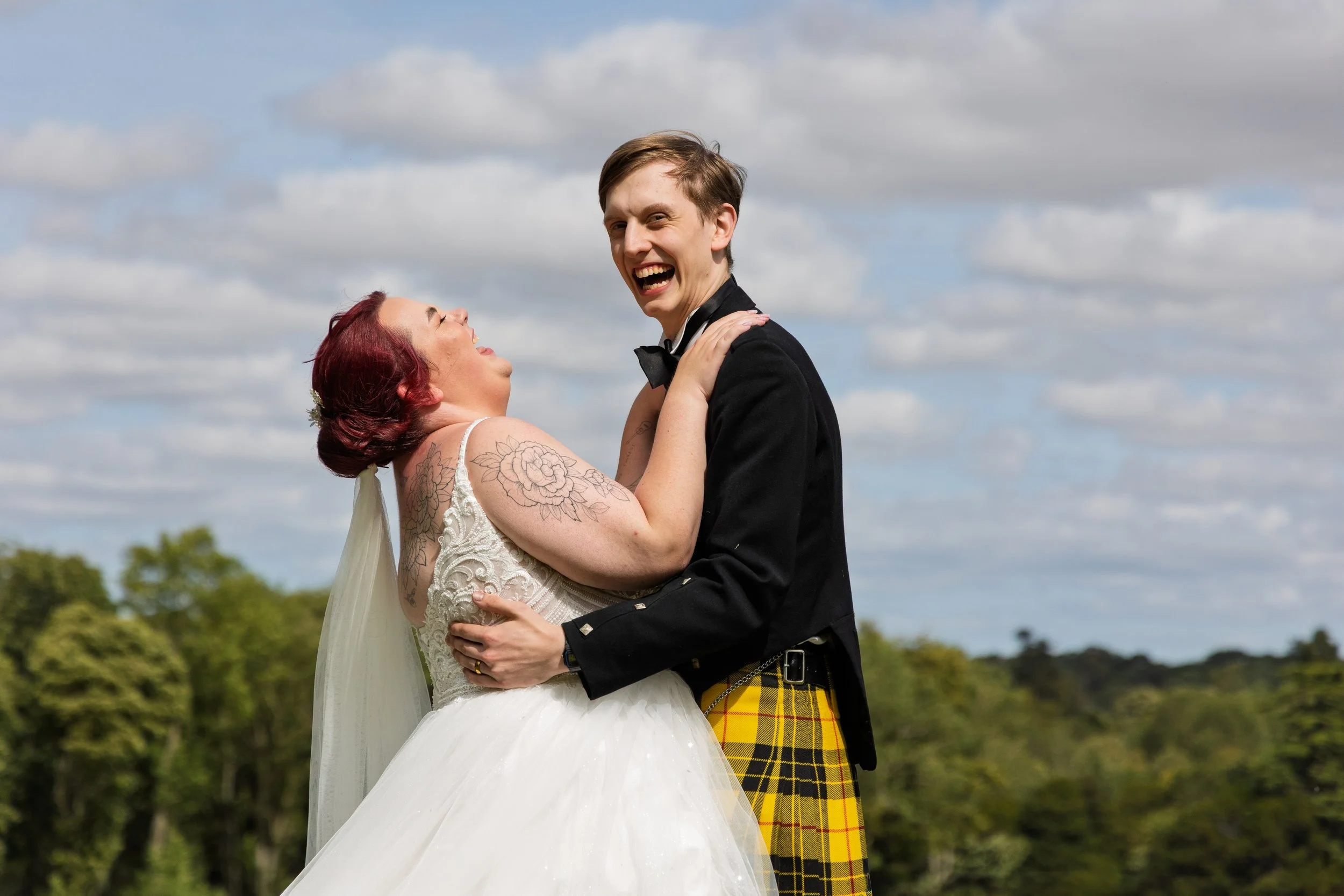 A bride and groom are dancing outside under a cloudy sky, smiling and looking at each other. The bride has red hair, tattoos on her arms, and is wearing a white wedding gown. The groom is wearing a black tuxedo with a bow tie and yellow plaid kilt.
