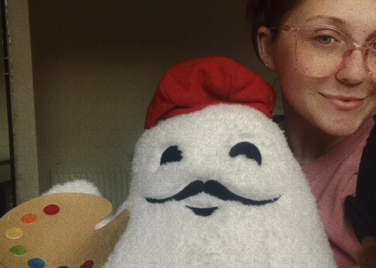 Part of a woman with glasses smiling next to a large fluffy plush toy resembling Santa Claus with a red hat, black mustache, and smiling face, holding a small artist's palette with colorful paint dots.