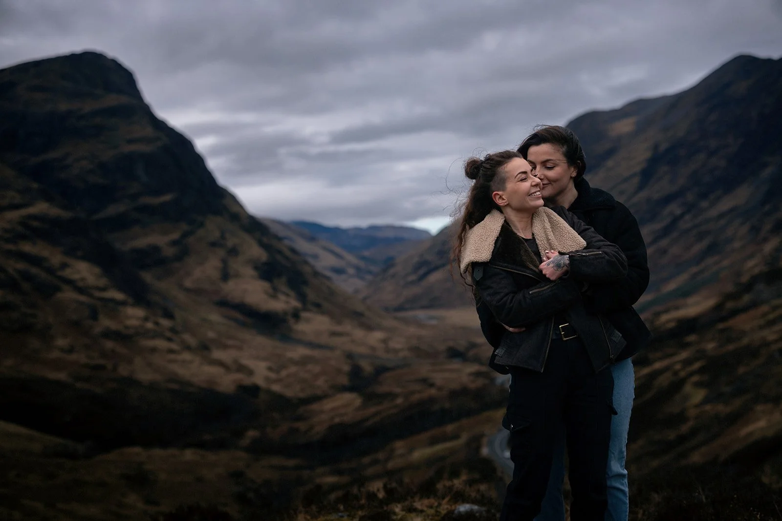 Two women hugging and smiling in a mountainous landscape with cloudy skies.