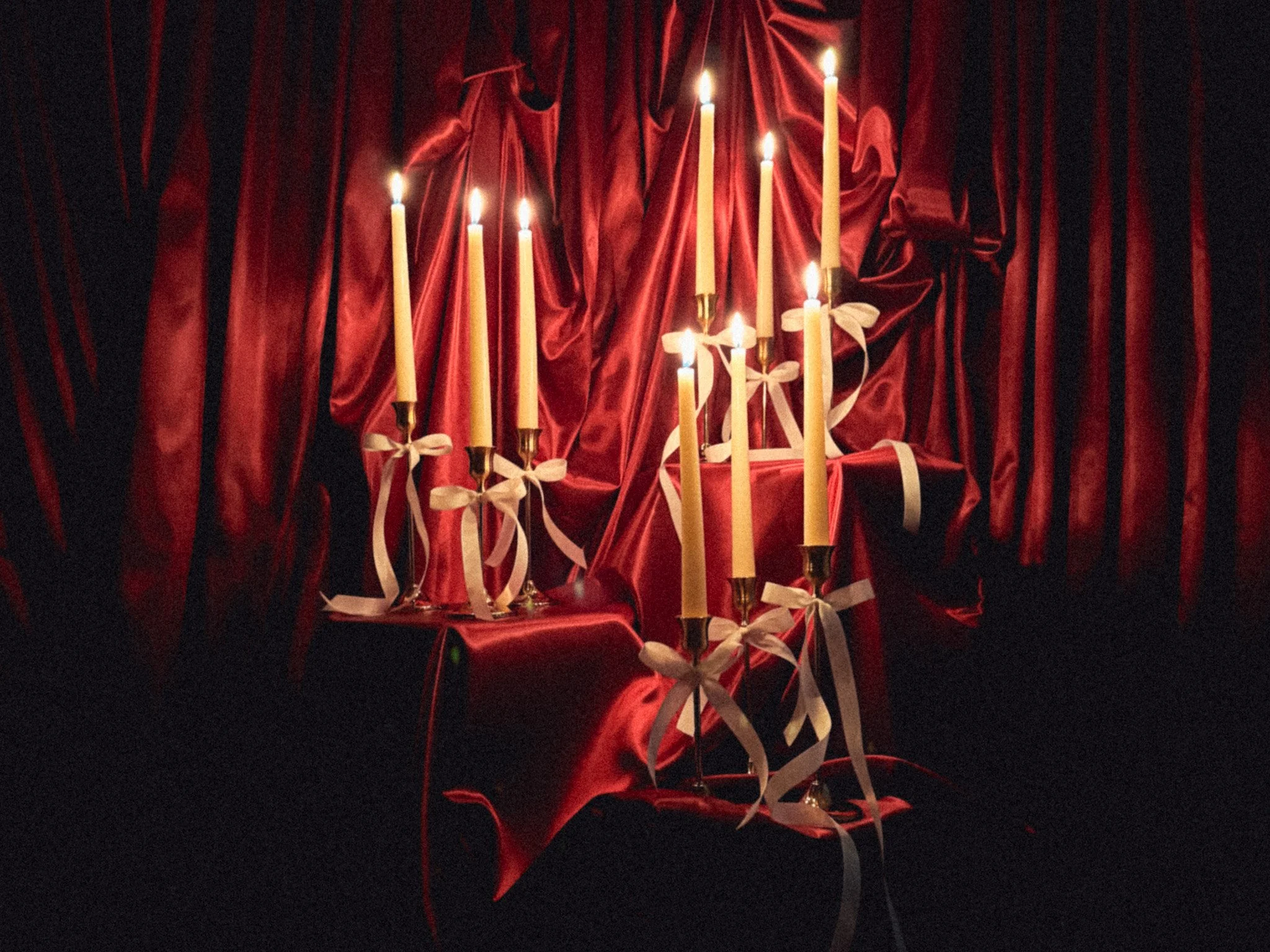 A candelabra with seven lit candles, draped with white ribbons, on a red satin-covered surface with a backdrop of red satin curtains.