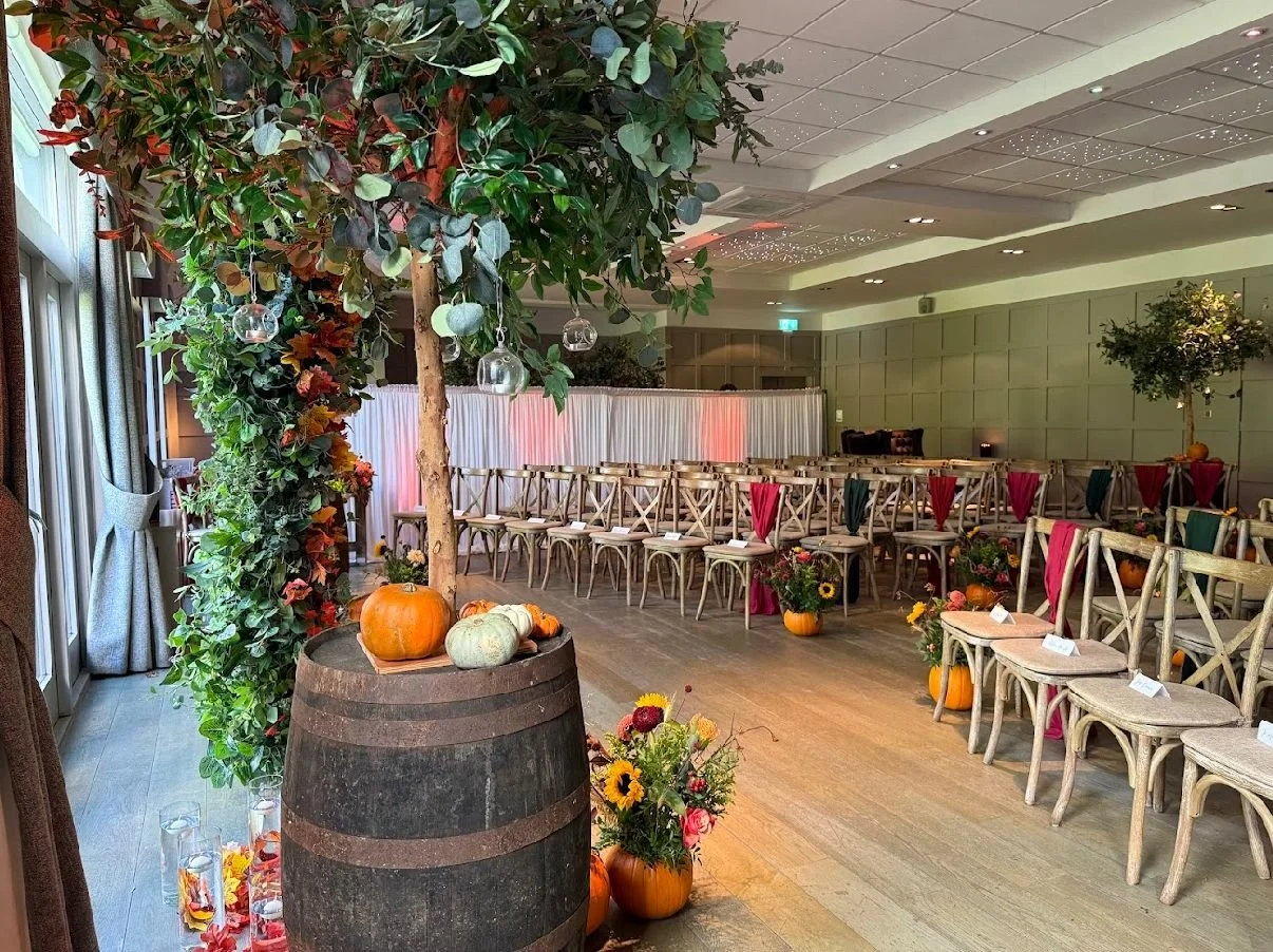 Decorated event space with wooden chairs, pumpkins, and floral arrangements, featuring a large tree with hanging glass ornaments and a barrel with pumpkins in the foreground.