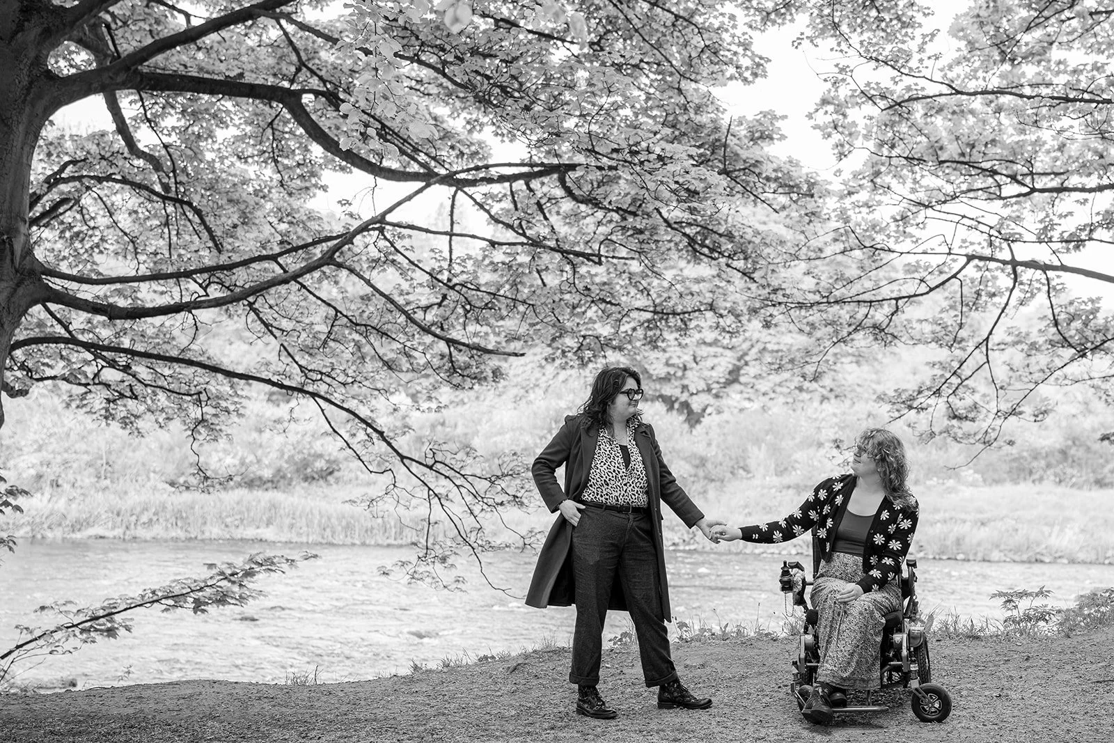 Two women outdoors near a river; one woman standing and holding the hand of the other woman who is seated in a motorized wheelchair. They are under a large tree with spreading branches and blooming leaves.