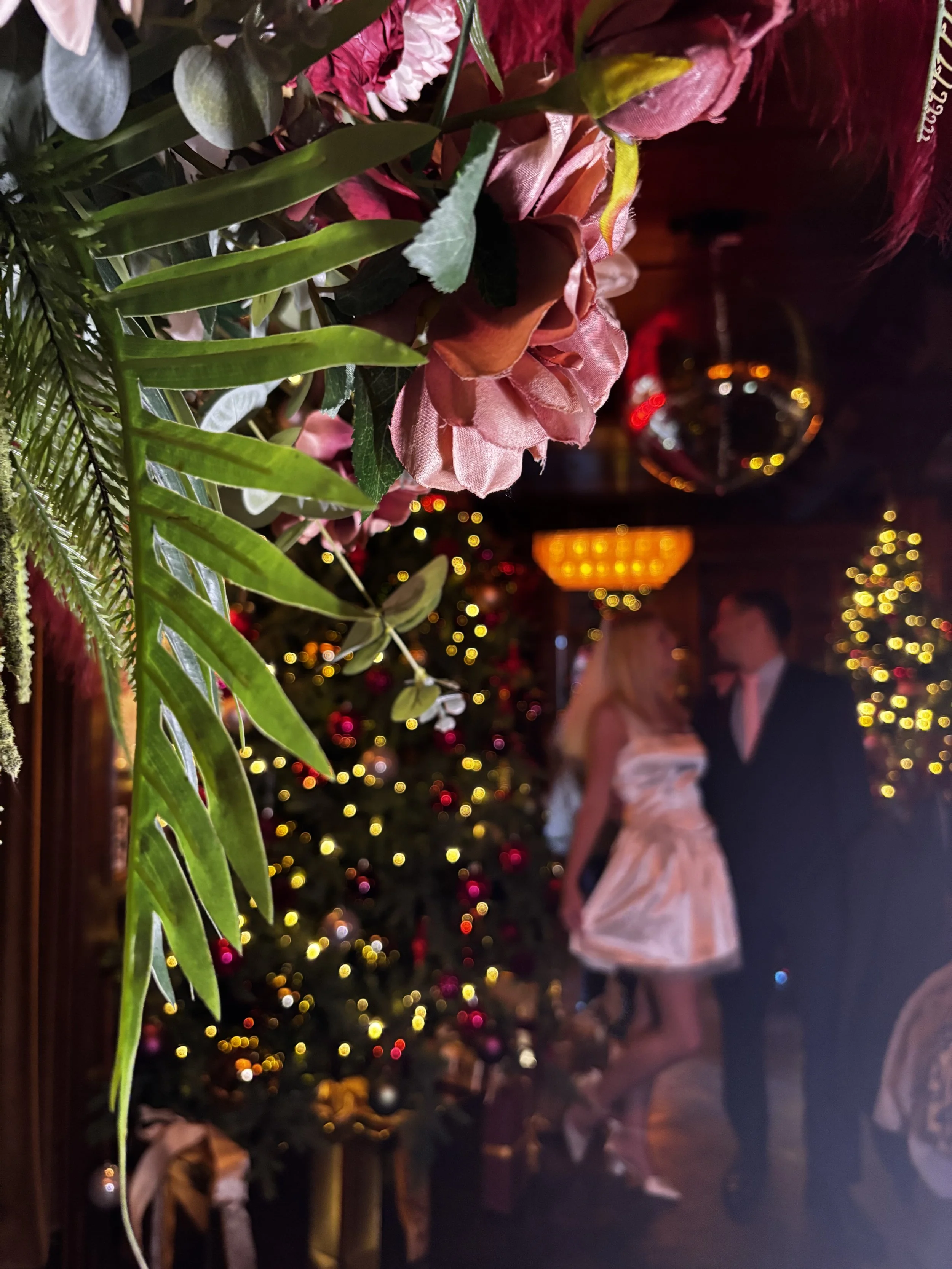 Close-up of pink and green flower arrangement with blurred background of Christmas decorated room, featuring a Christmas tree with lights and ornaments, a couple dressed formally standing and talking, festive lights, and a disco ball.