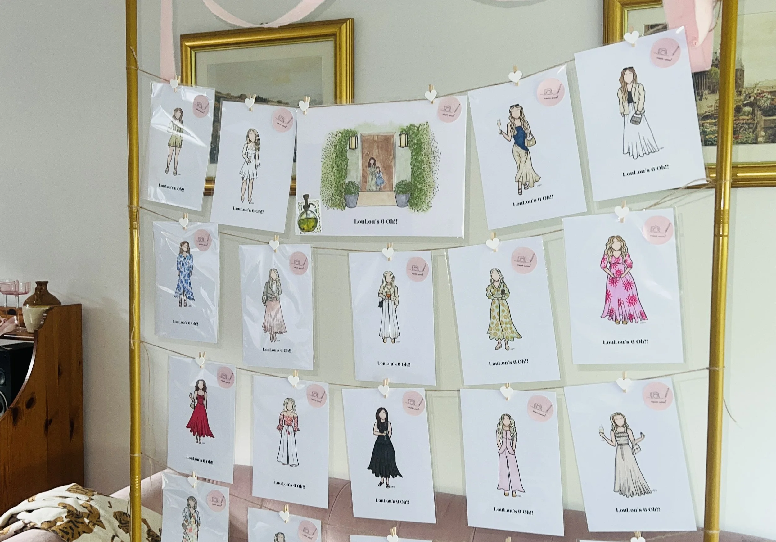 Colorful drawings of women in various outfits hanging on a yellow frame with pink heart-shaped clips, with some drawings in plastic covers and a small container of perfume on the display.