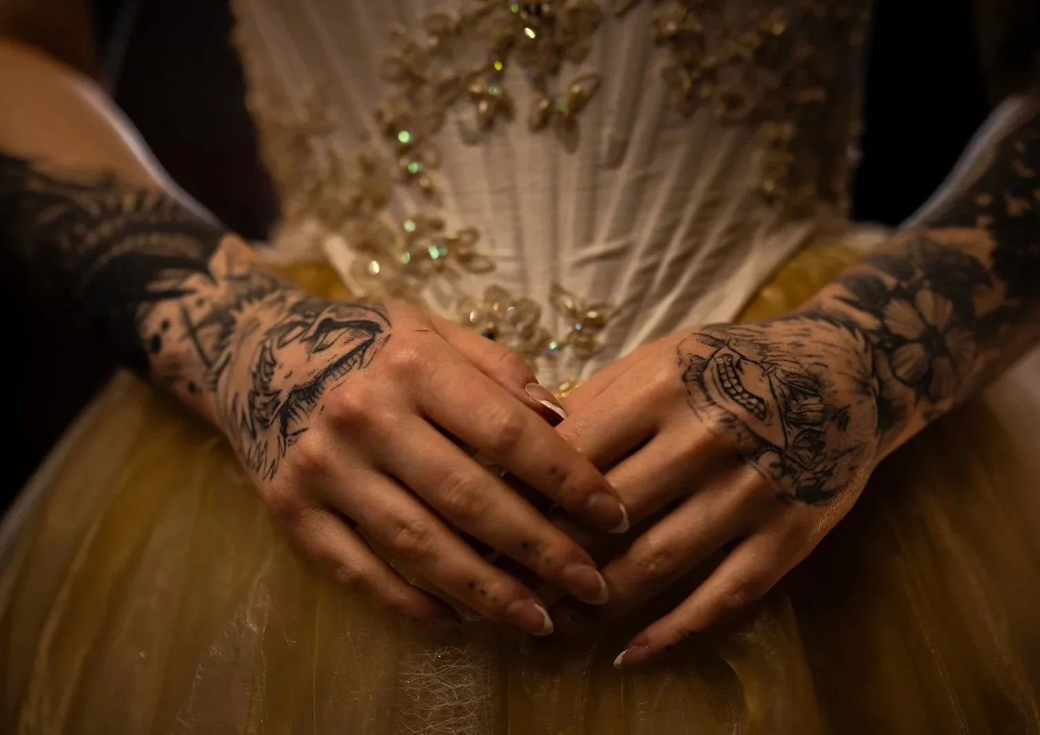 Close-up of a person with elaborate tattoos on both hands, sitting with hands clasped on lap, wearing a detailed outfit with embroidery and sequins.