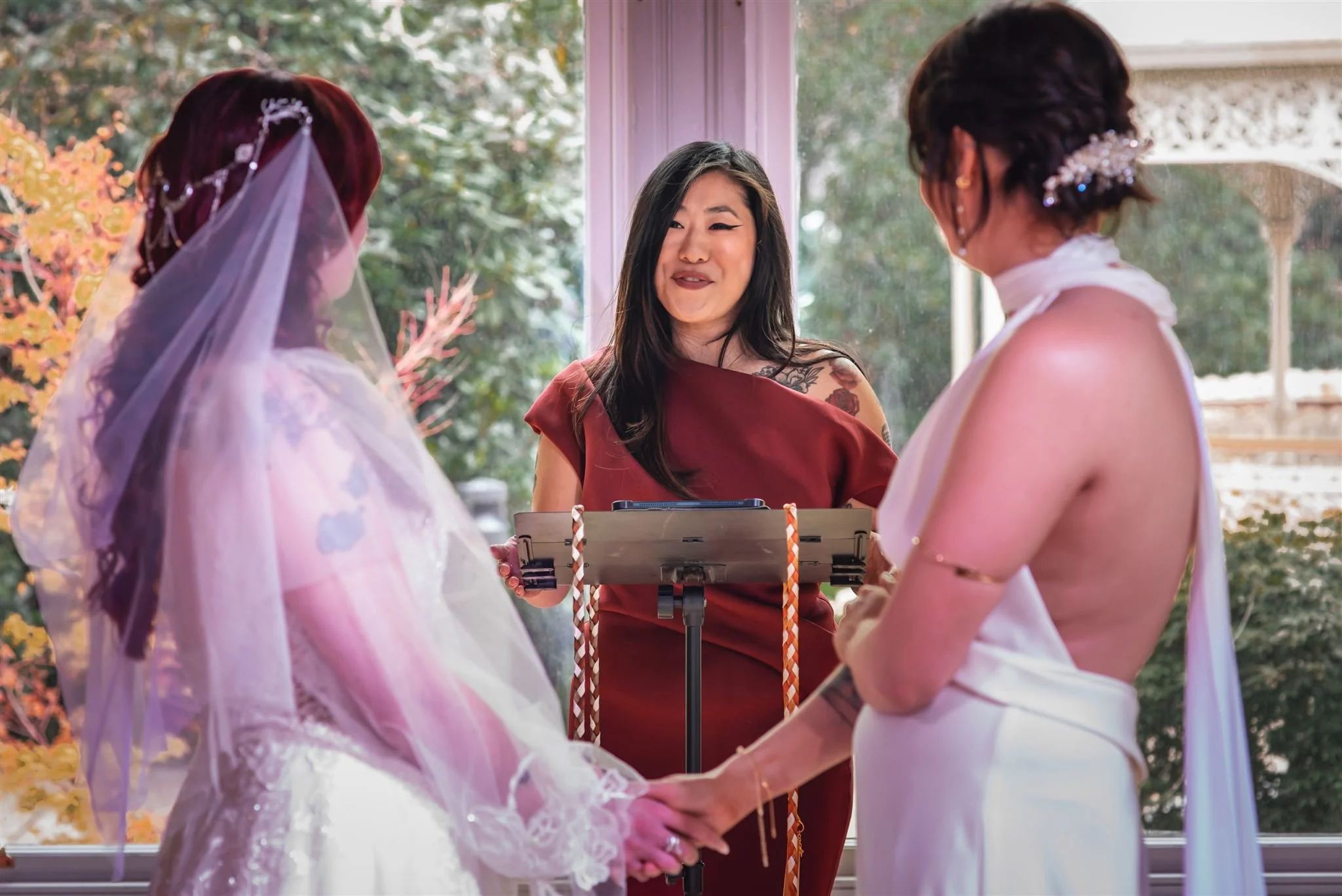Two women standing facing each other holding hands, with a third woman officiating a wedding ceremony in front of them, inside a room with large windows showing greenery outside.