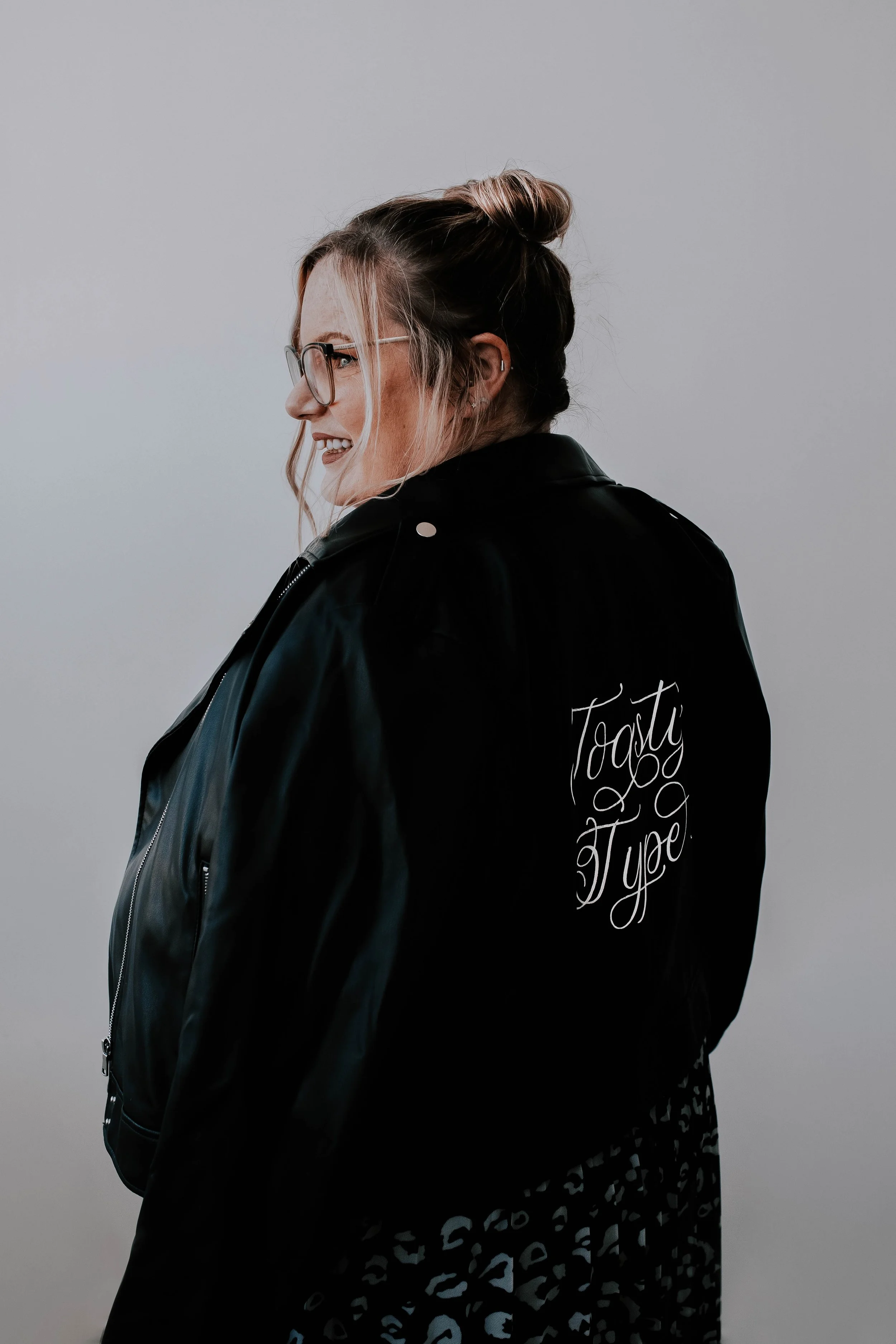A woman with glasses and a messy bun hairstyle, wearing a black jacket with 'Tasty Type' written on the back, standing against a plain grey background.