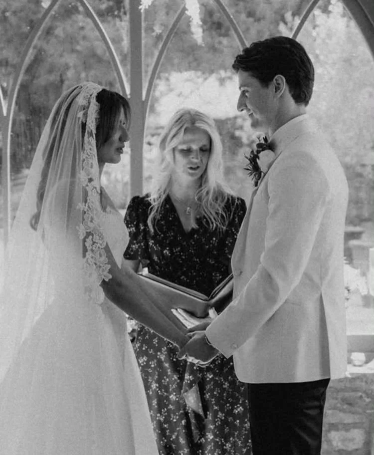 A black and white photo of a wedding ceremony, with a bride and groom holding hands and facing each other, an officiant reading from a book between them, set in a church or bridal venue.