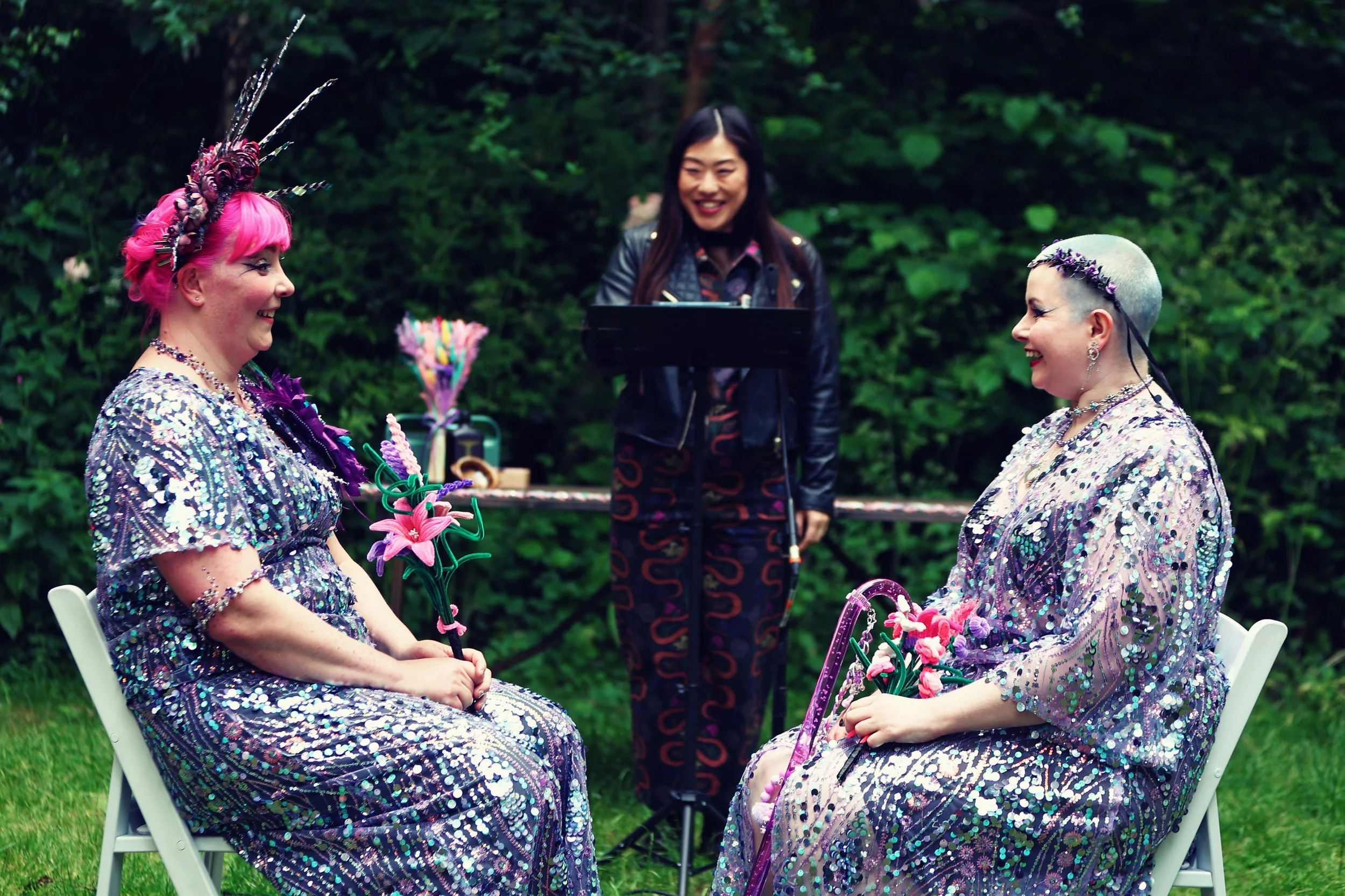Two women dressed in shiny, colorful, sequin outfits sit facing each other, holding flower-shaped objects. They are smiling at each other with an officiant standing behind them, outdoors, with green foliage in the background.