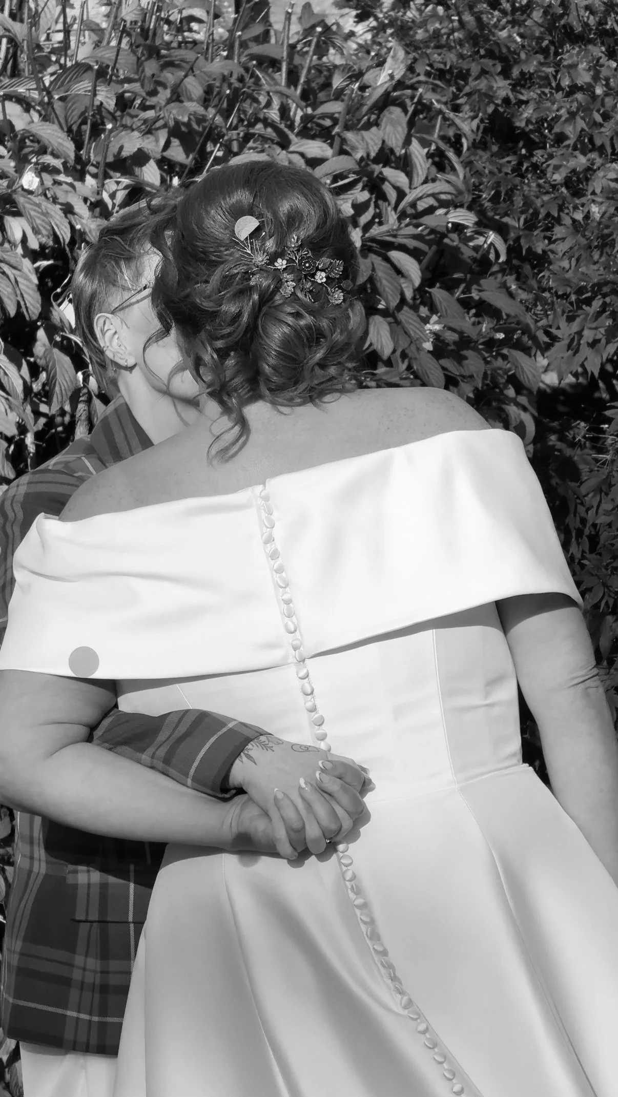 A bride in a white off-the-shoulder wedding dress with button detail on the back, holding hands with a partner, in an outdoor setting with leafy background.