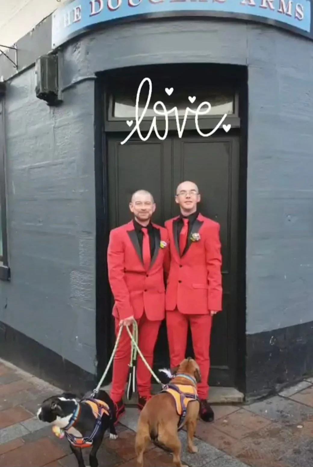 Two men in matching red suits with black lapels and ties standing in front of a black door, each holding a leash attached to a small dog wearing harnesses. The photo has the word 'love' written above them with small heart doodles. The scene appears to be outside a building with gray walls and a brick sidewalk.