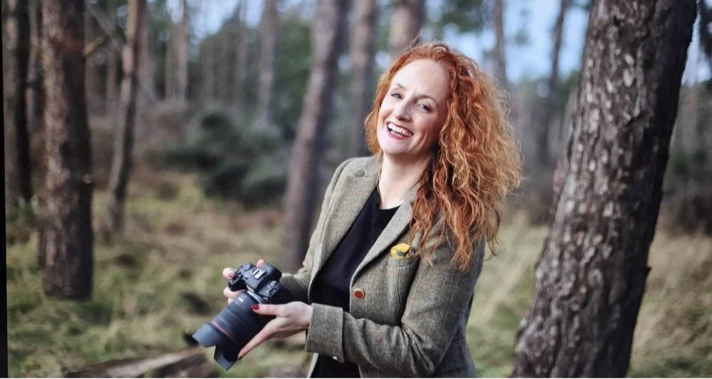 A woman with red curly hair smiling and holding a camera in a forest.