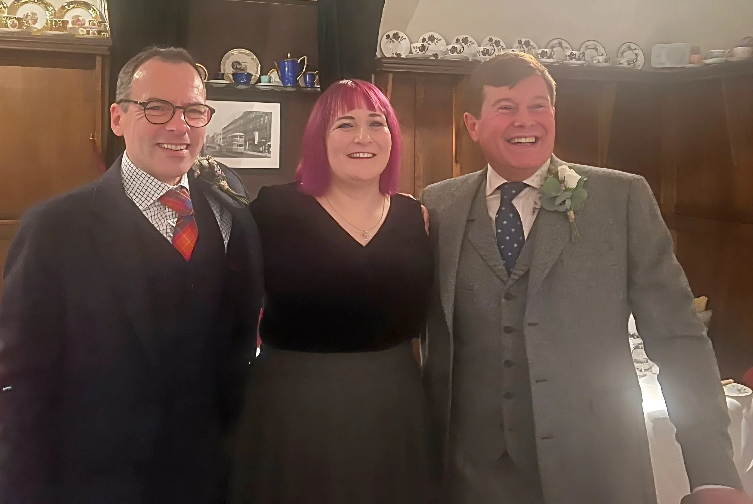 Three people standing together, smiling, inside a room with dark wooden cabinets and decorative plates displayed on top. One man is wearing glasses and a suit with a checkered shirt and a red tie, the woman in the middle has pink hair and is wearing a black top, and the other man is in a gray suit with a dark tie and a boutonniere.