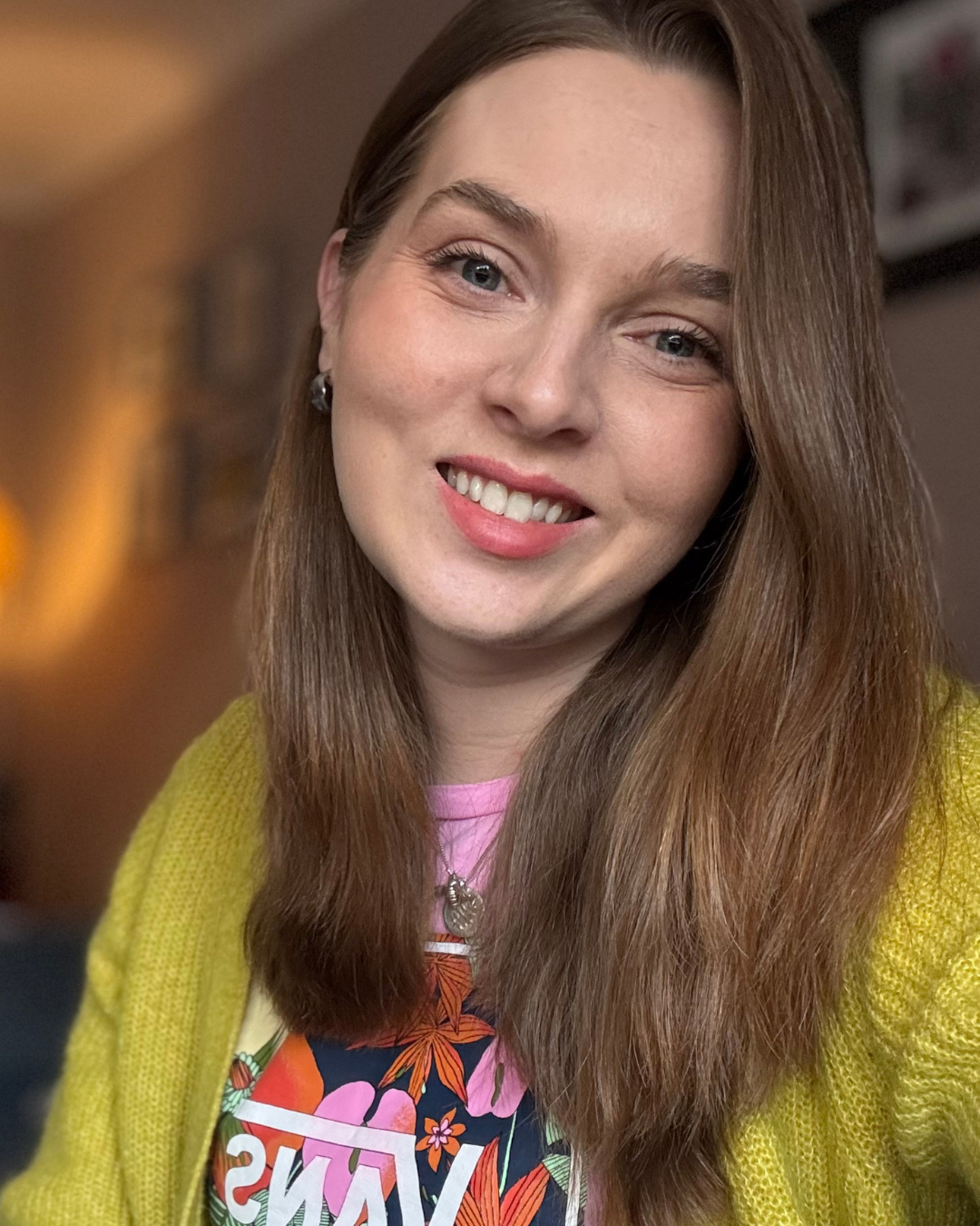 Close-up of a smiling woman with long brown hair, wearing a yellow cardigan over a colorful floral shirt, in an indoor setting.