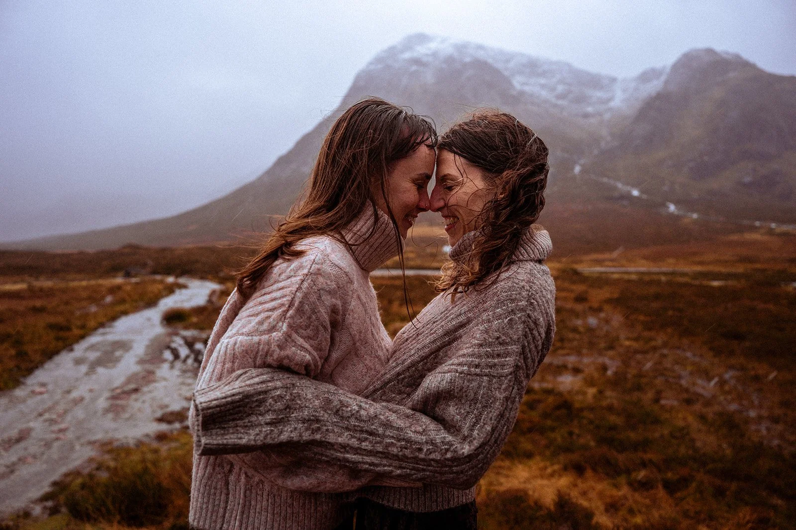Two women, wearing cozy sweaters, embrace with foreheads touching, smiling at each other in a misty mountain landscape with a dirt trail and snow-capped peaks in the background.