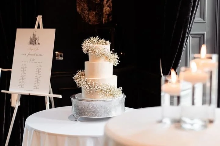 Three-tier white wedding cake with baby's breath flowers on a silver cake stand, decorated with white fondant, on a white tablecloth.