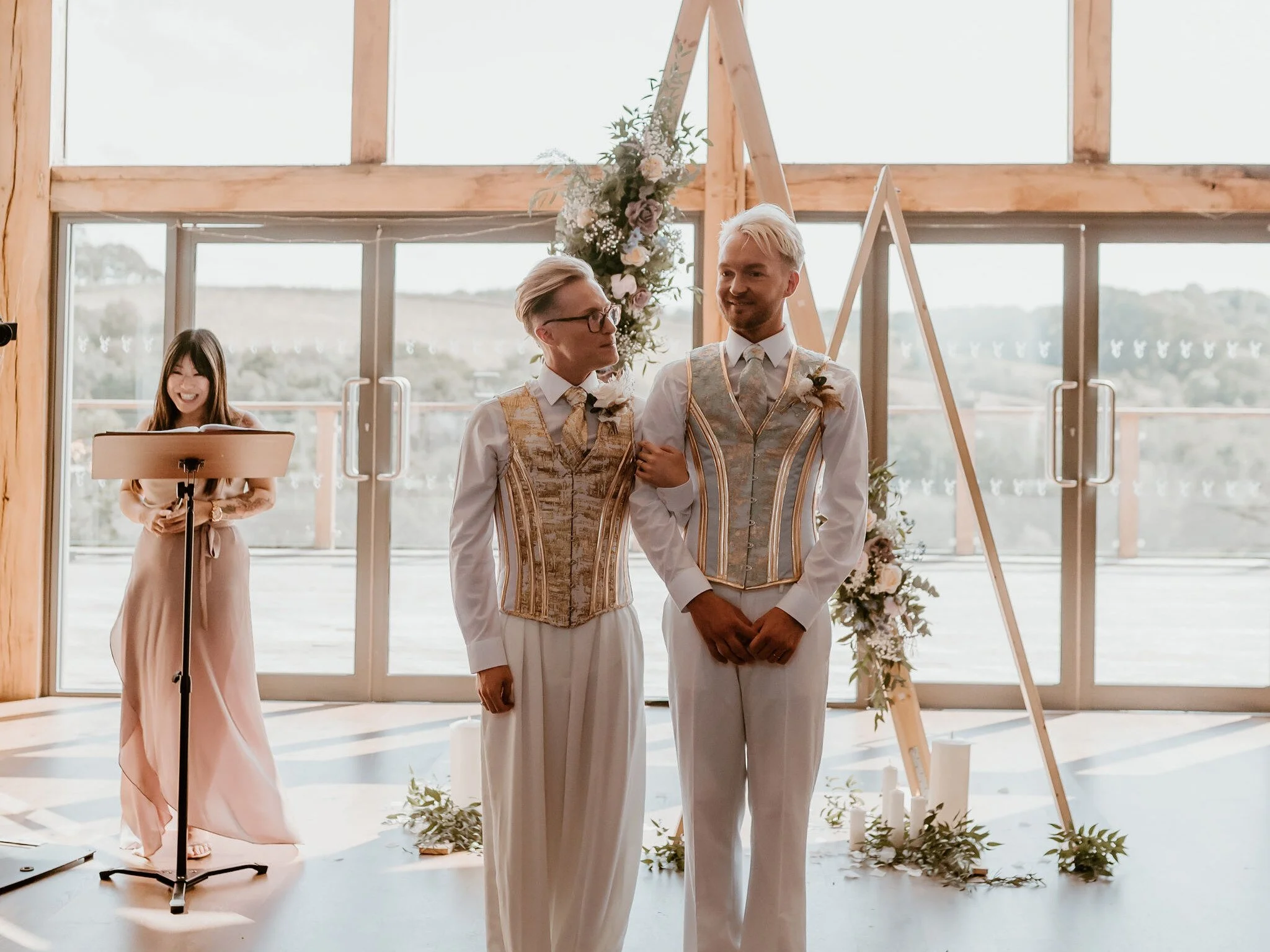 Two grooms holding hands and smiling during their wedding ceremony, with a bridesmaid standing nearby, in front of large windows with a scenic outdoor view.