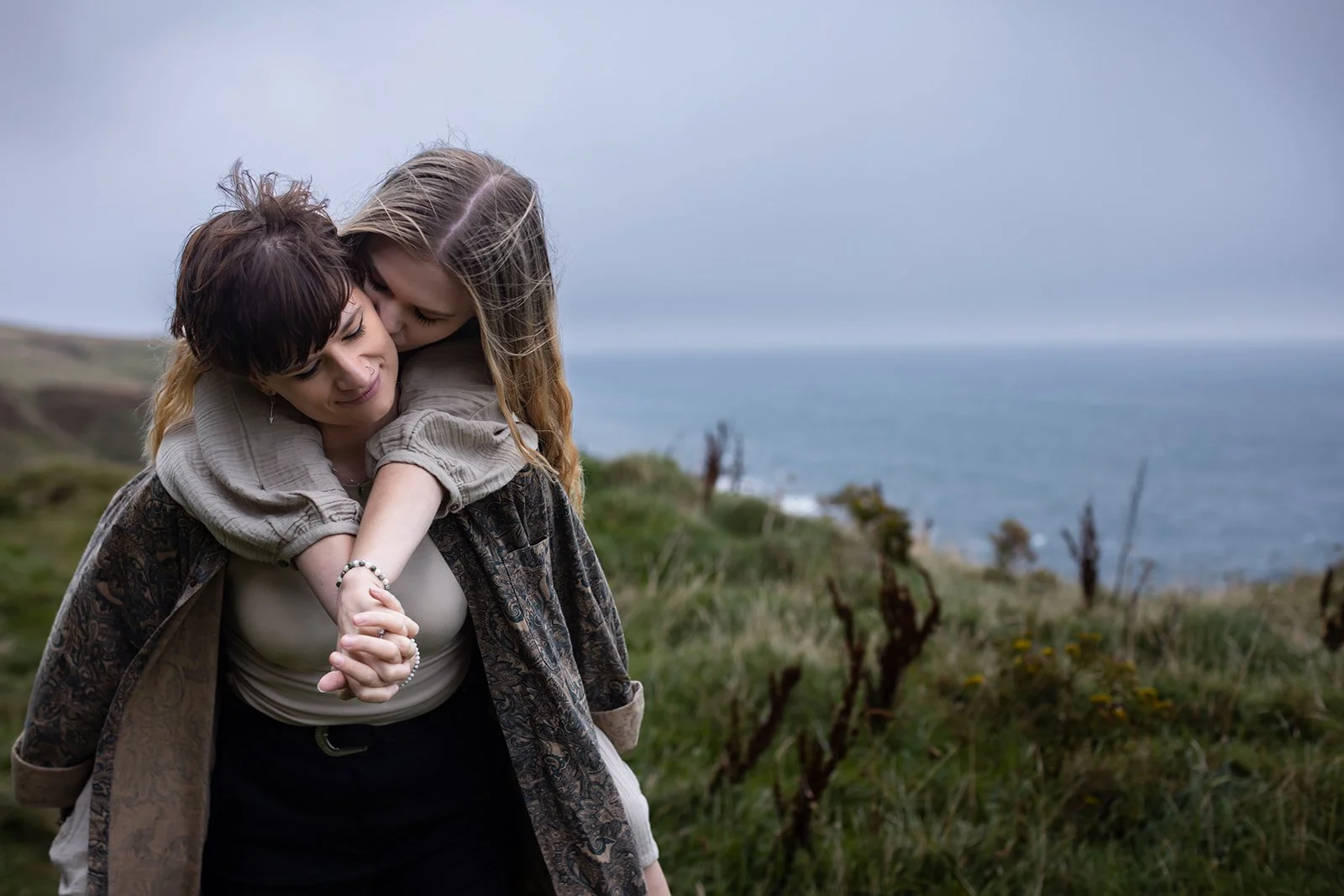 A woman is carrying a young girl on her back, and they are hugging each other outdoors near the coast with grassy hills and a cloudy sky in the background.