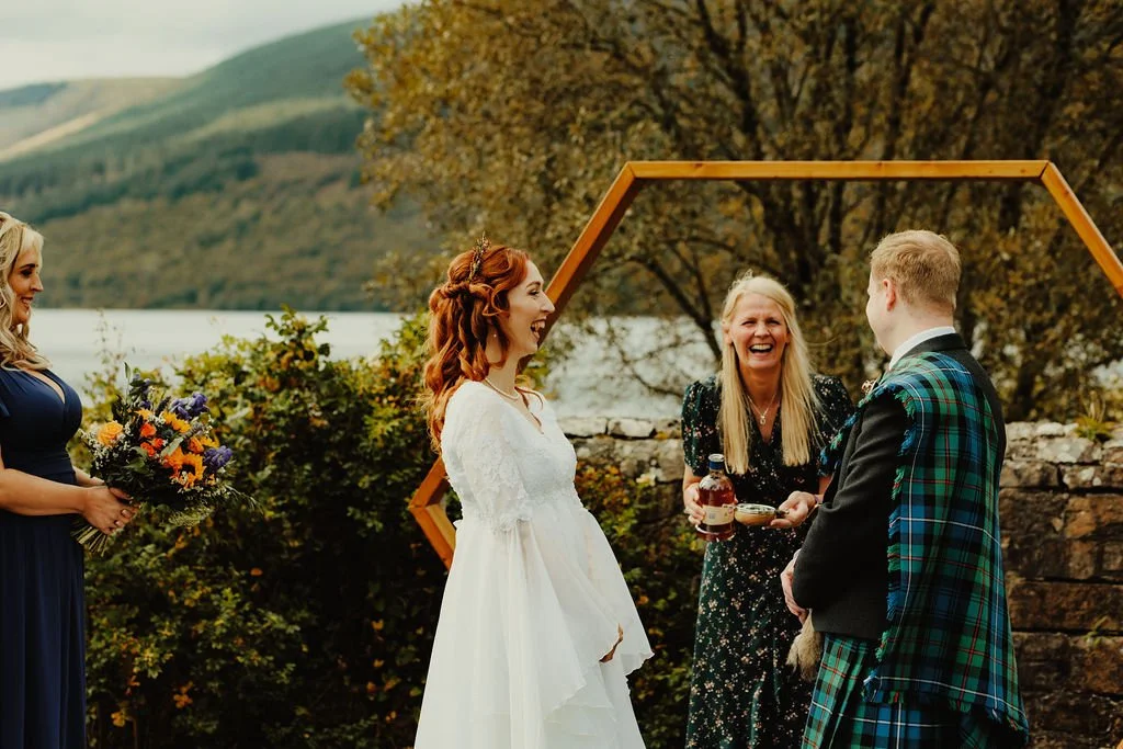 A wedding ceremony outdoors with a bride in a white dress, a groom in a tartan kilt, a woman officiant, and a bridesmaid holding a bouquet. They are smiling with a scenic lake and mountains in the background.