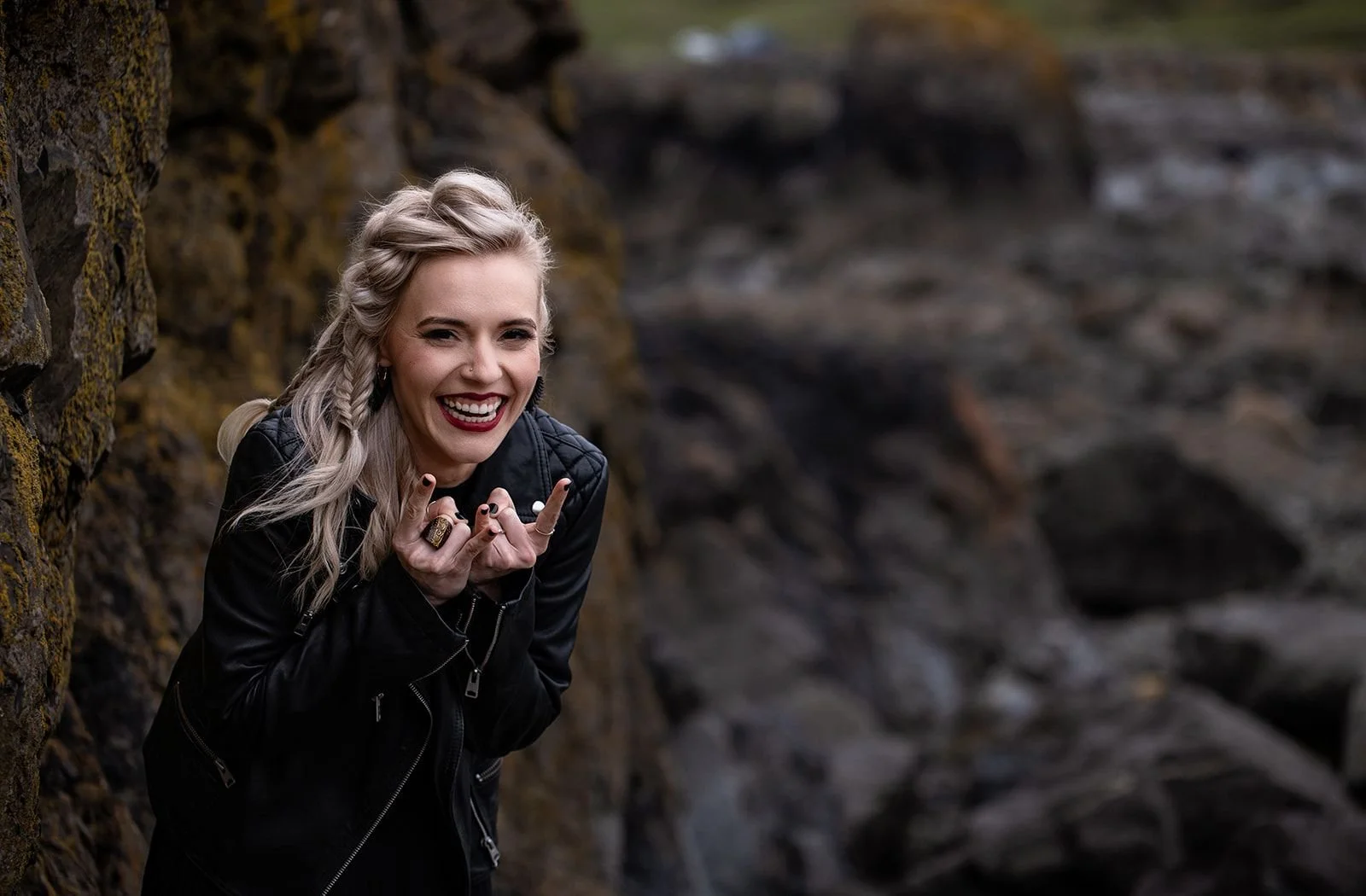 A smiling woman with long, blonde hair styled in braids, wearing a black leather jacket, making a rock and roll hand gesture with both hands in an outdoor setting near rocks or a cliff.