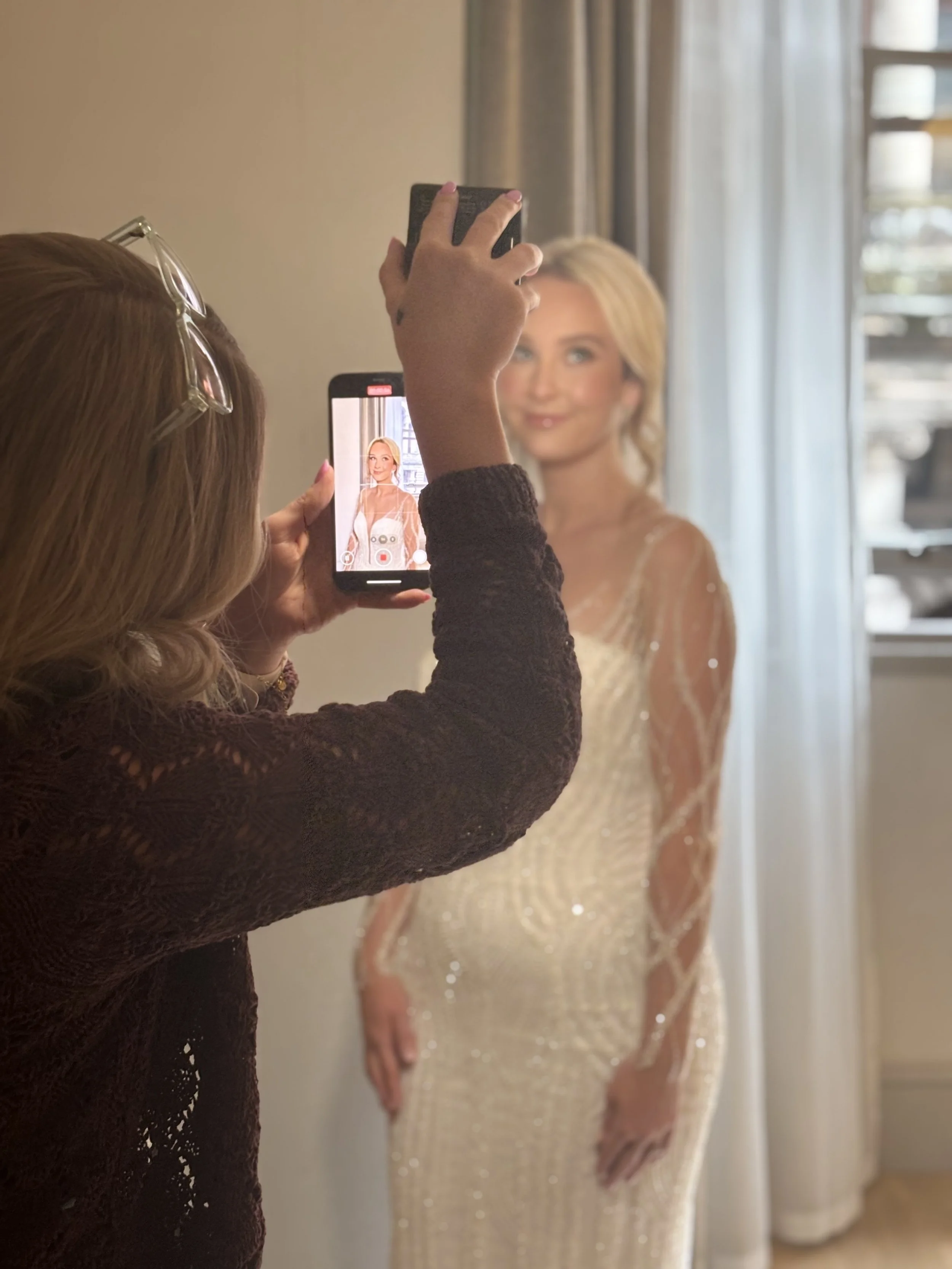 A woman takes a photo of another woman in a sparkling gown using her smartphone in front of a mirror.