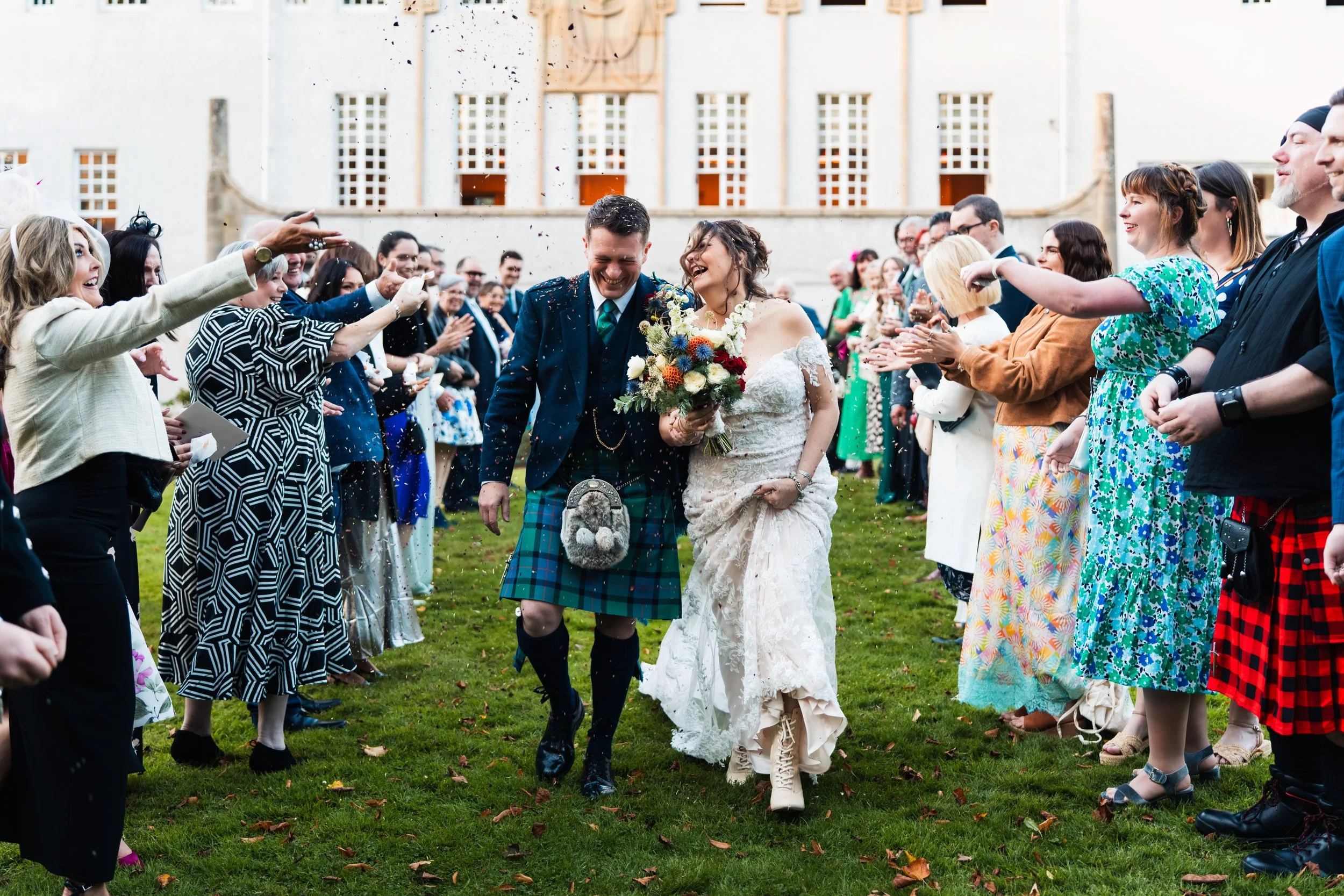 Wedding celebration with a bride and groom walking through a crowd of friends and family on a green lawn, outdoors in front of a large building, with guests cheering and throwing confetti.