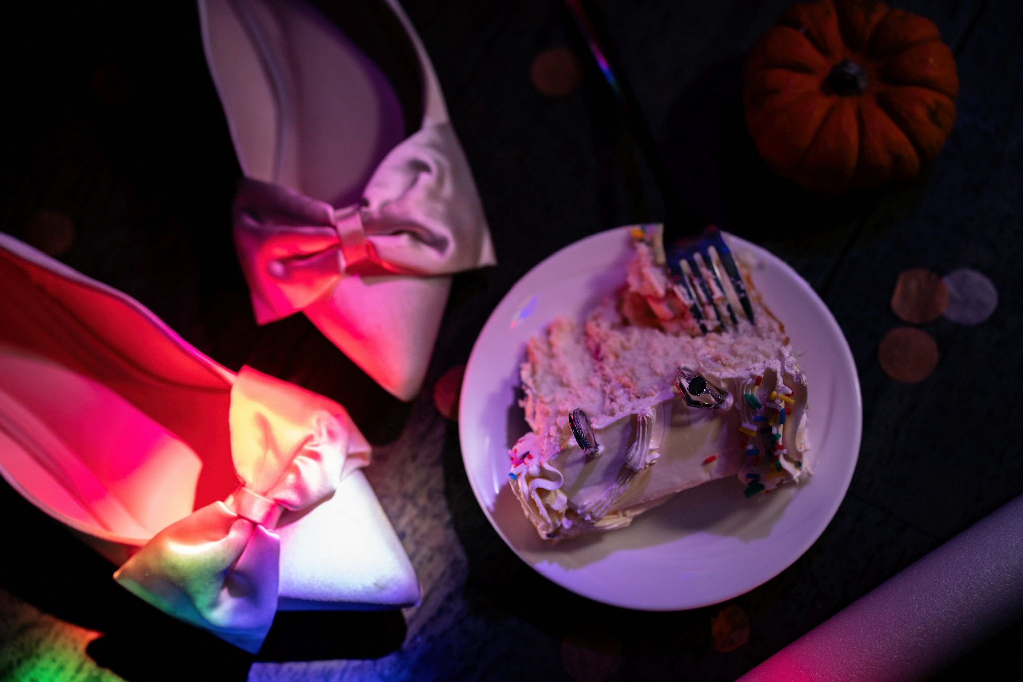 A slice of cake with white frosting and colorful sprinkles on a white plate, surrounded by rainbow-colored napkins tied with pink ribbons, a small pumpkin, and scattered autumn leaves.