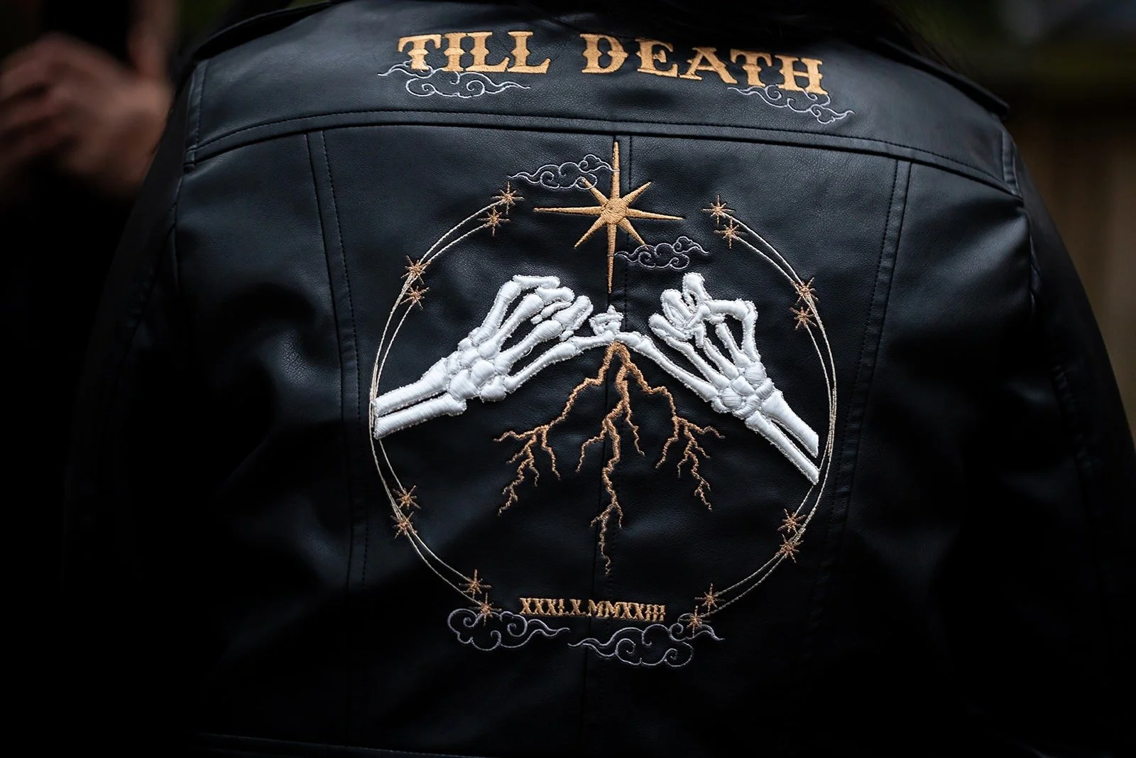 Back of a black leather jacket with embroidered designs including skeletal hands, a star, clouds, lightning bolts, and the text 'TILL DEATH' at the top and 'XXXL MCMXVI' at the bottom.