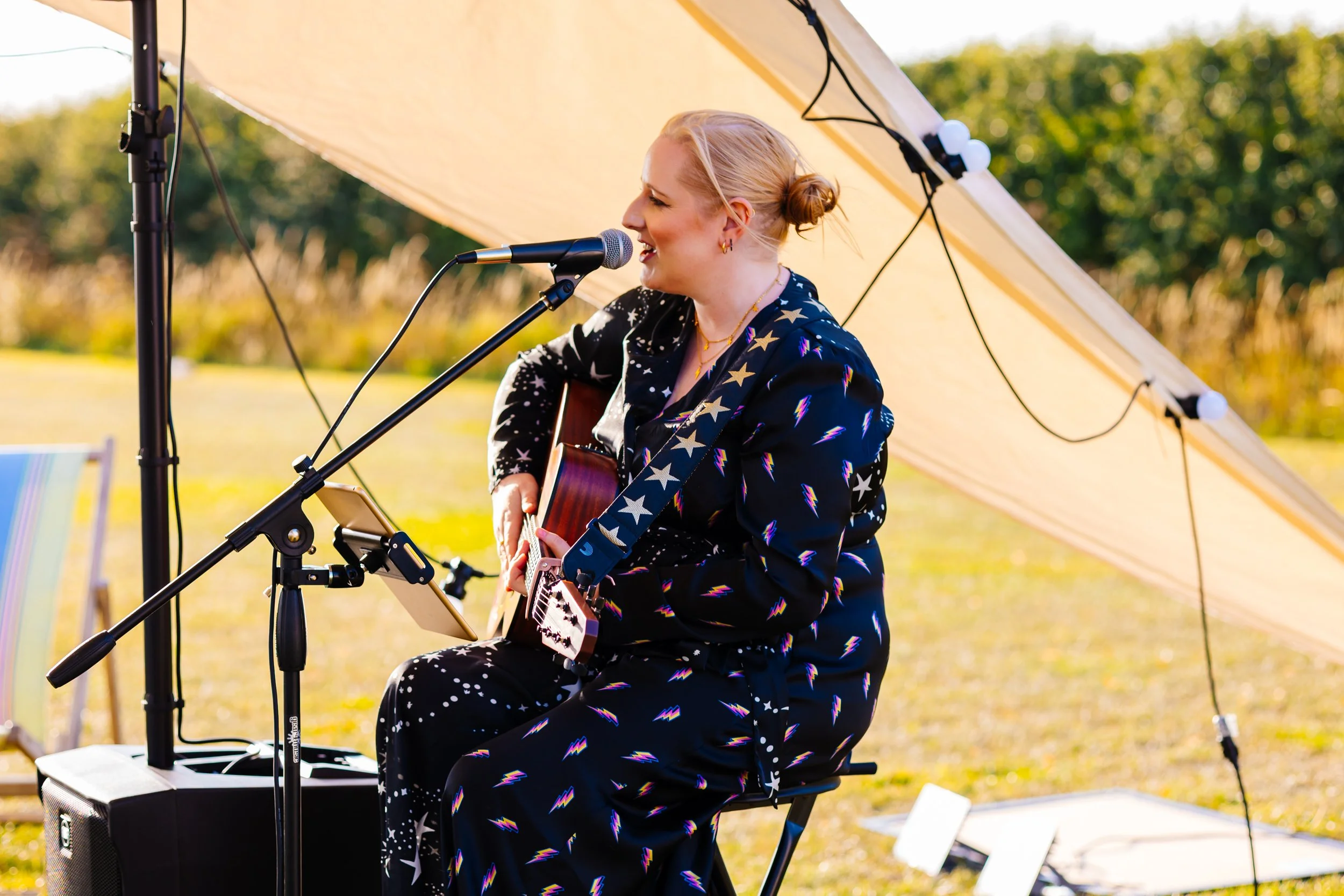 A woman with blonde hair tied in a bun, wearing a star and lightning bolt patterned black outfit, is playing an acoustic guitar and singing into a microphone outdoors under a canopy on a sunny day.