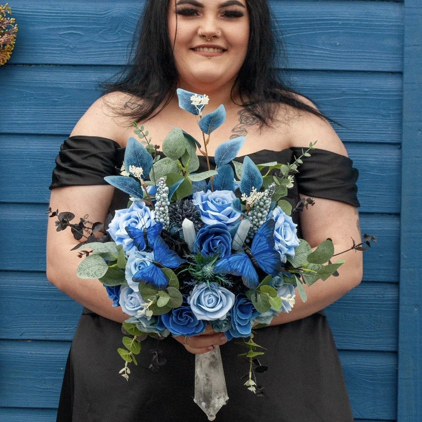 Woman with dark hair and makeup holding a bouquet of blue roses, butterflies, and greenery standing in front of a blue wooden wall.