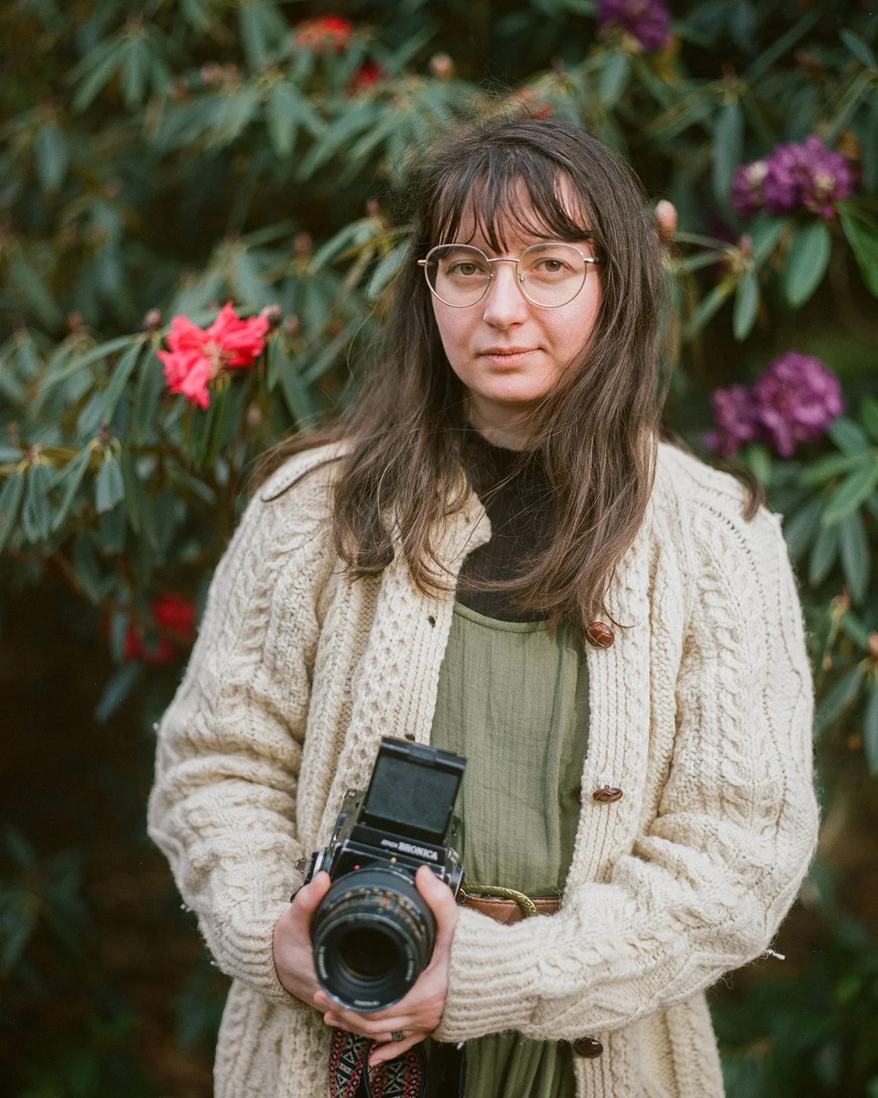 A woman with long brown hair and glasses holding a vintage camera in front of green foliage and colorful flowers.